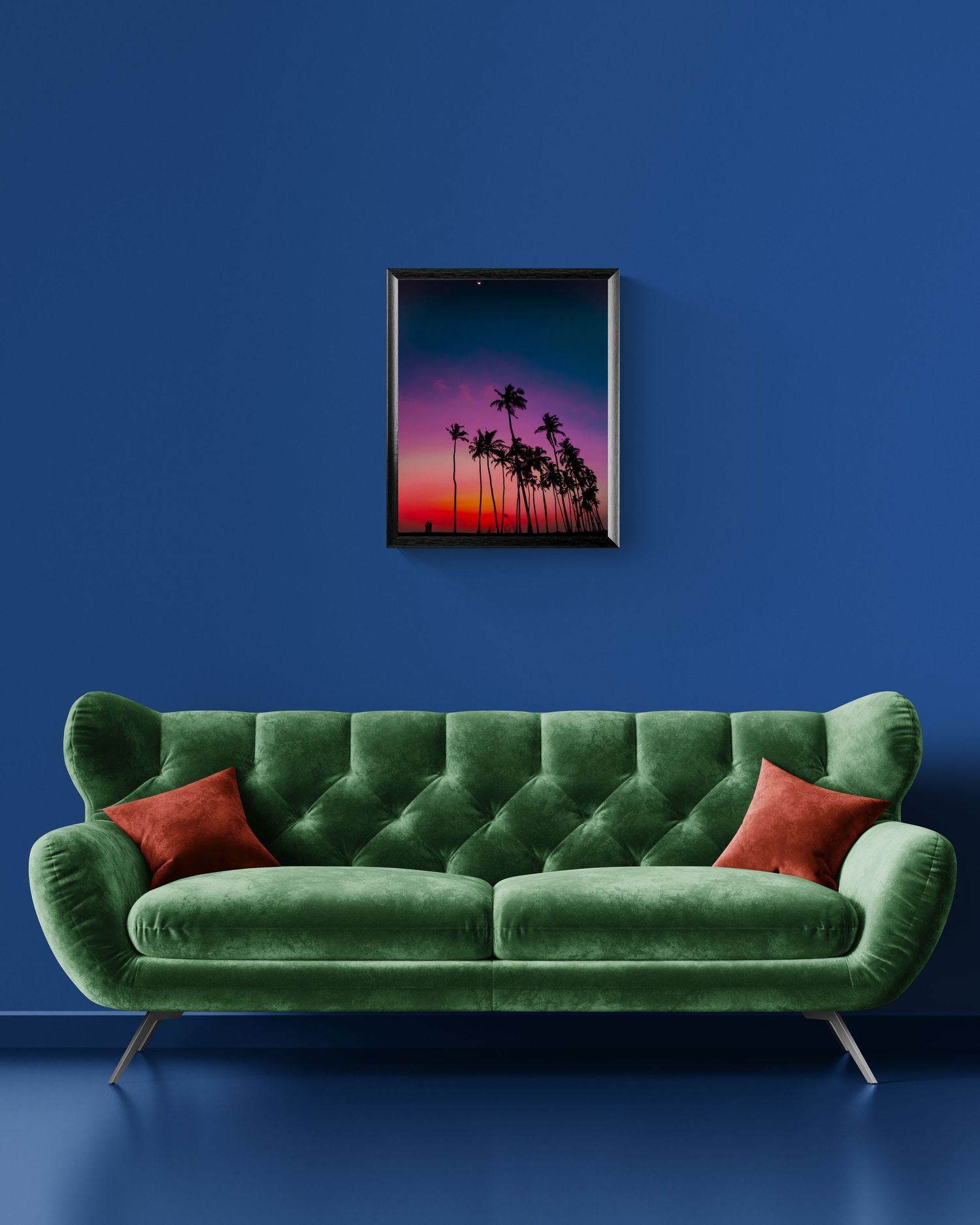Green velvet tufted sofa with red pillows against a cobalt blue wall, beneath a vibrant tropical sunset palm tree framed print.
