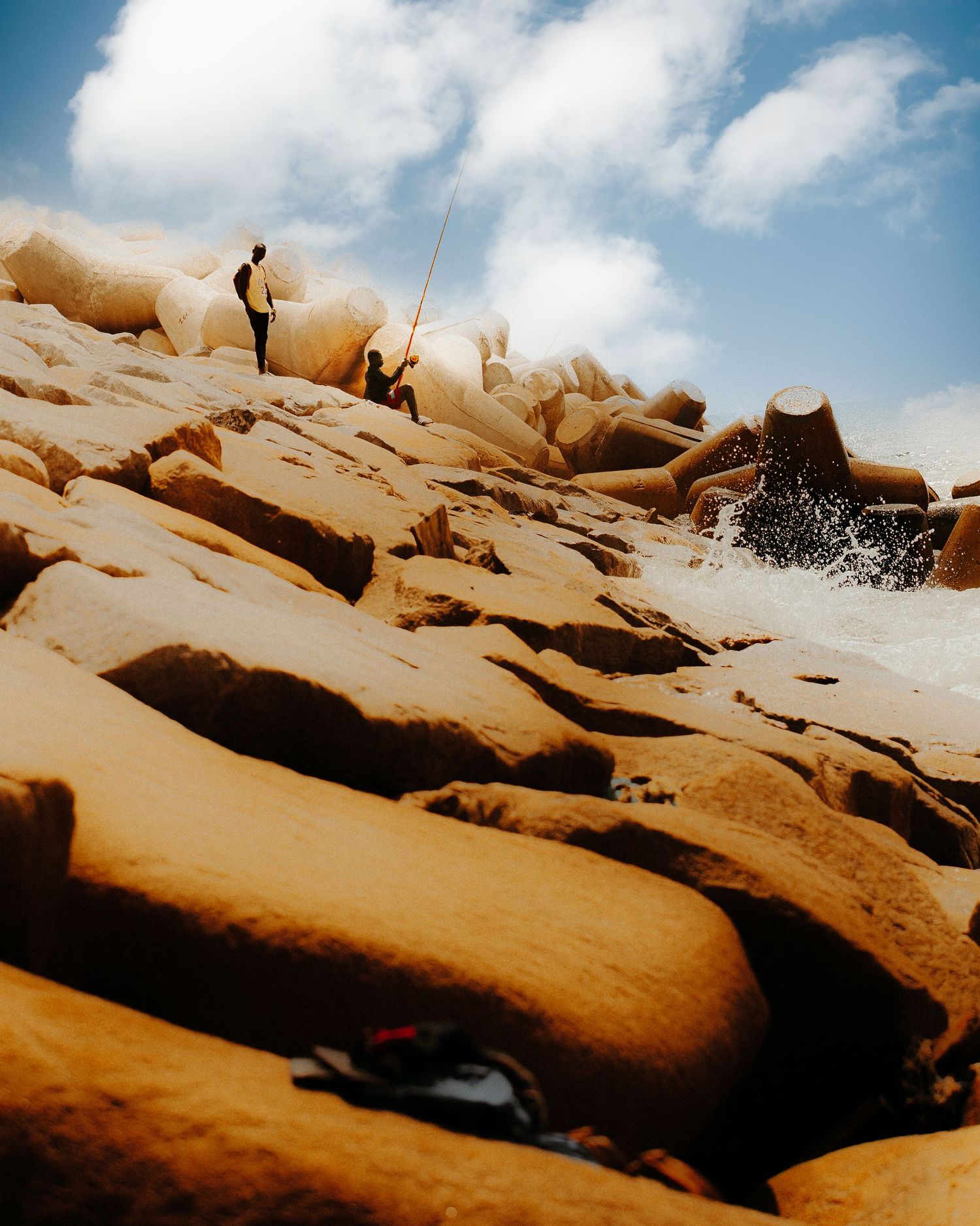 Person fishing on rugged golden rocks by crashing ocean waves under a dramatic cloudy sky.
