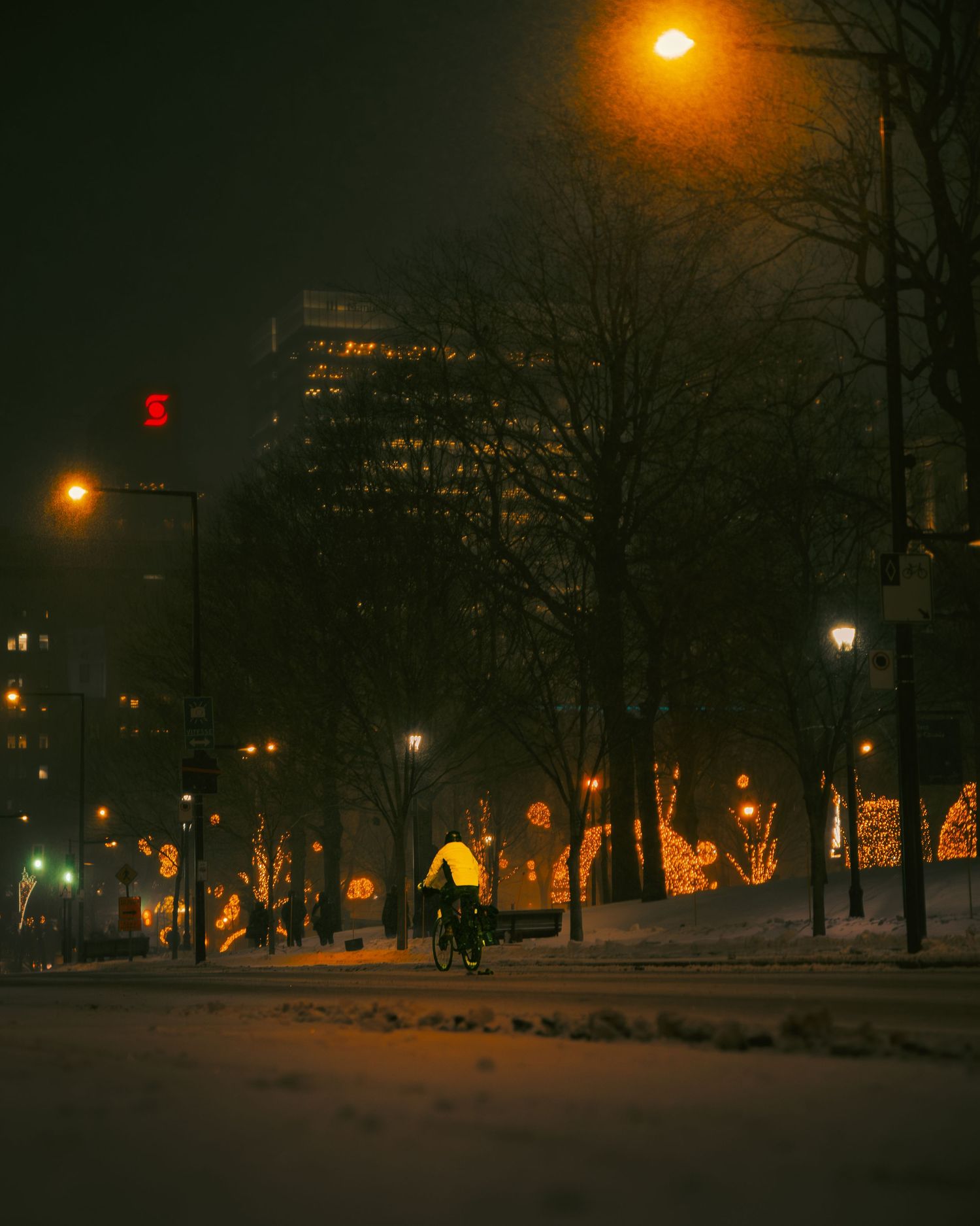 A cyclist rides through a snowy city street at night, illuminated by warm street lights and festive holiday decorations.