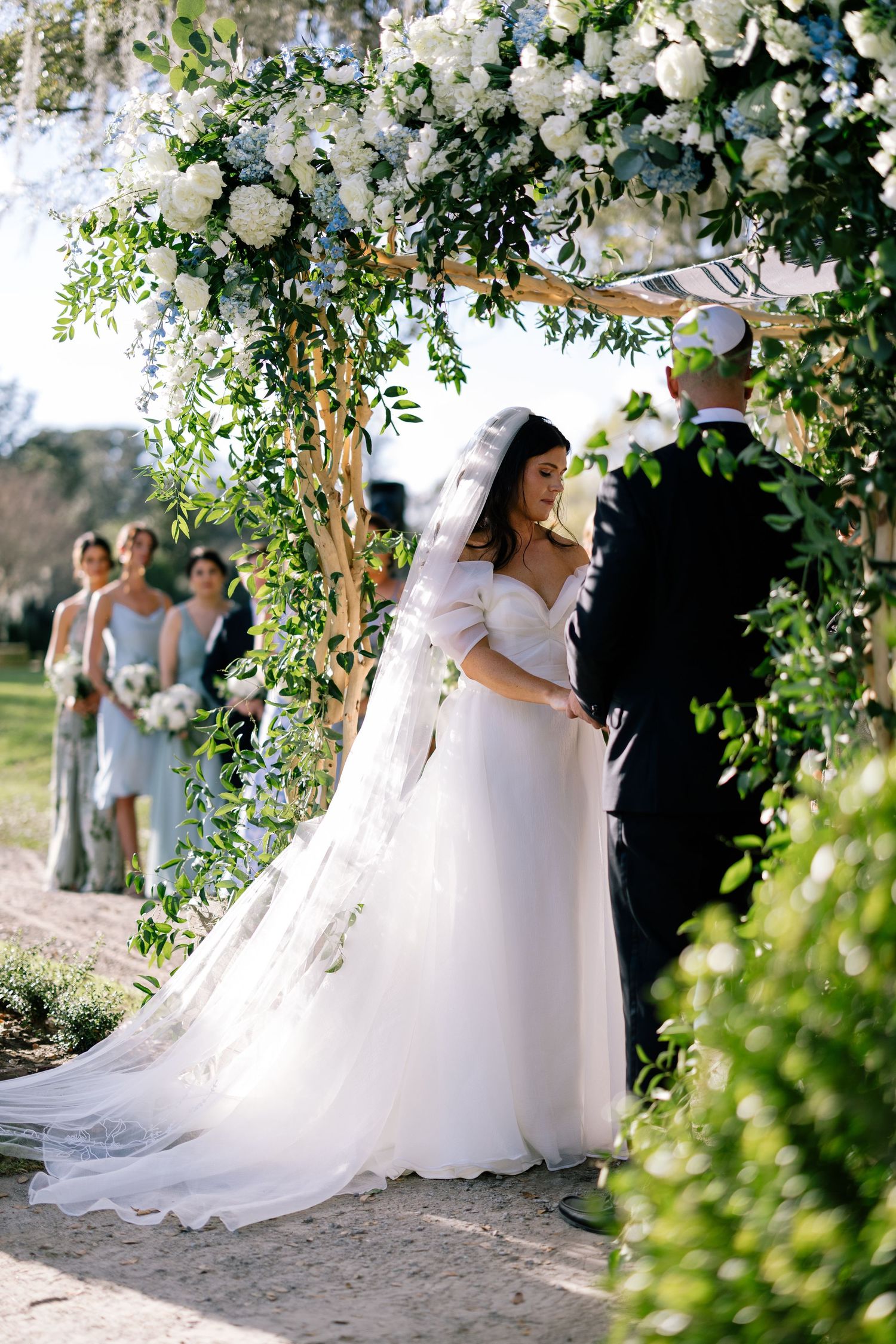 A bride and groom standing under a stunning blue floral display and chuppah. Bridesmaids line up in the background with blue dresses.