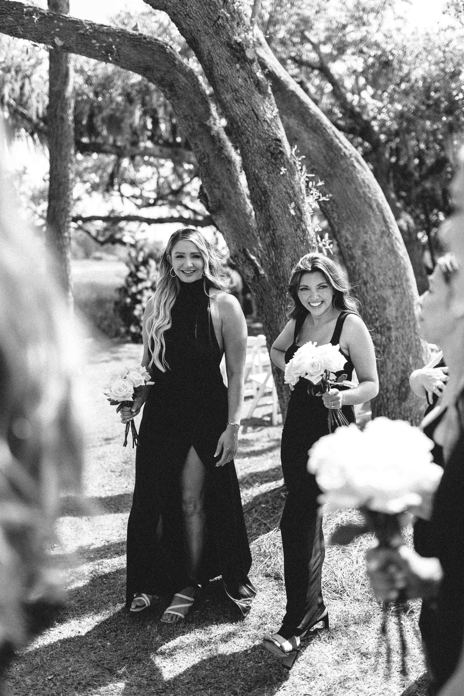 Black and white photo of two bridesmaids in black dresses holding bouquets, smiling while standing under trees during the ceremony