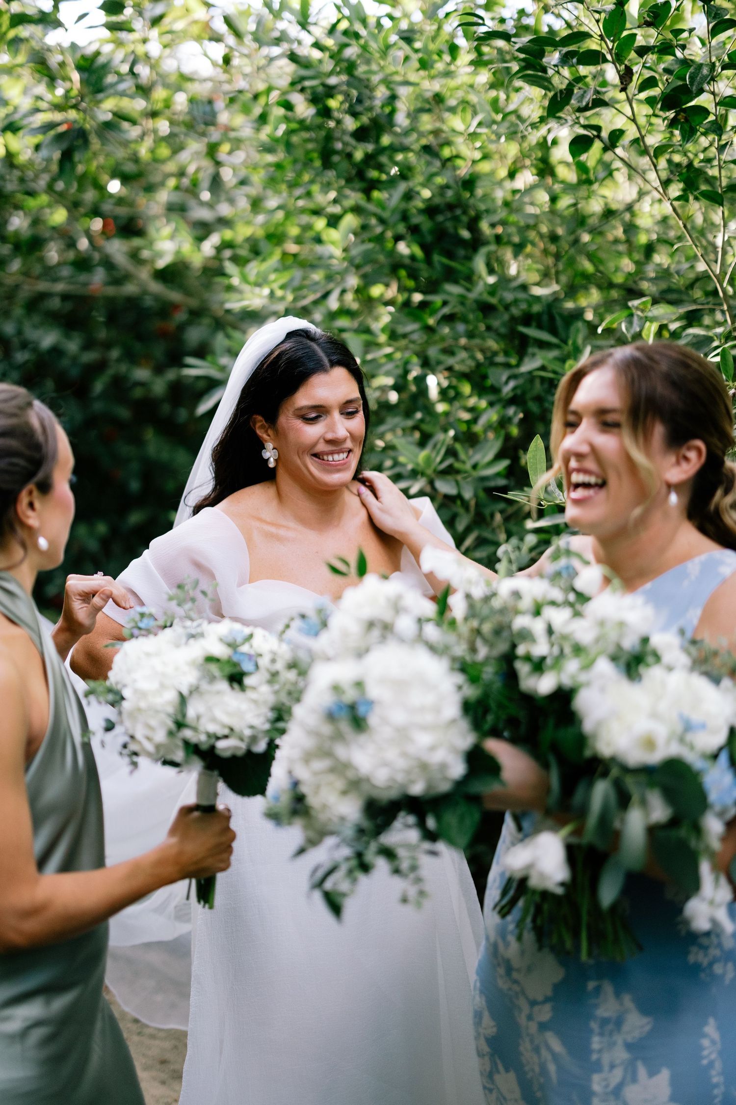 a bride smiling candidly with her bridesmaids holding wedding flowers, in a garden with tall shrubs
