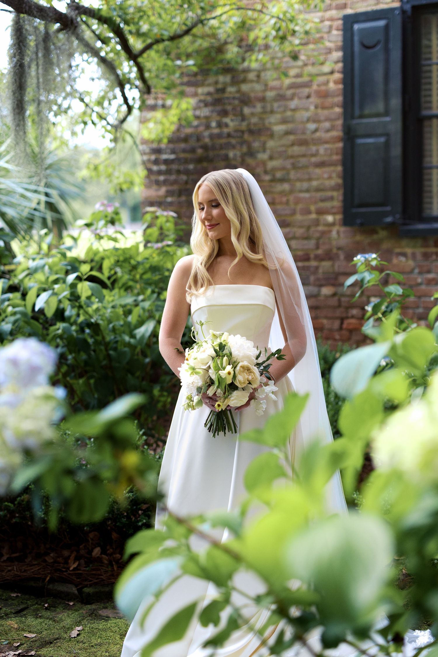 Bride in a strapless white gown and veil holding a bouquet of white and blush flowers, standing gracefully in a garden at Middleton Place