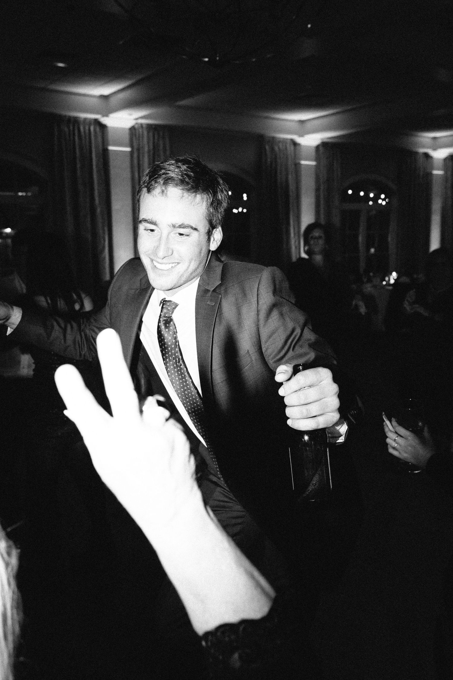 A black and white image of a smiling groom dancing at a wedding reception while holding a beer bottle. A peace sign signed by another guest.