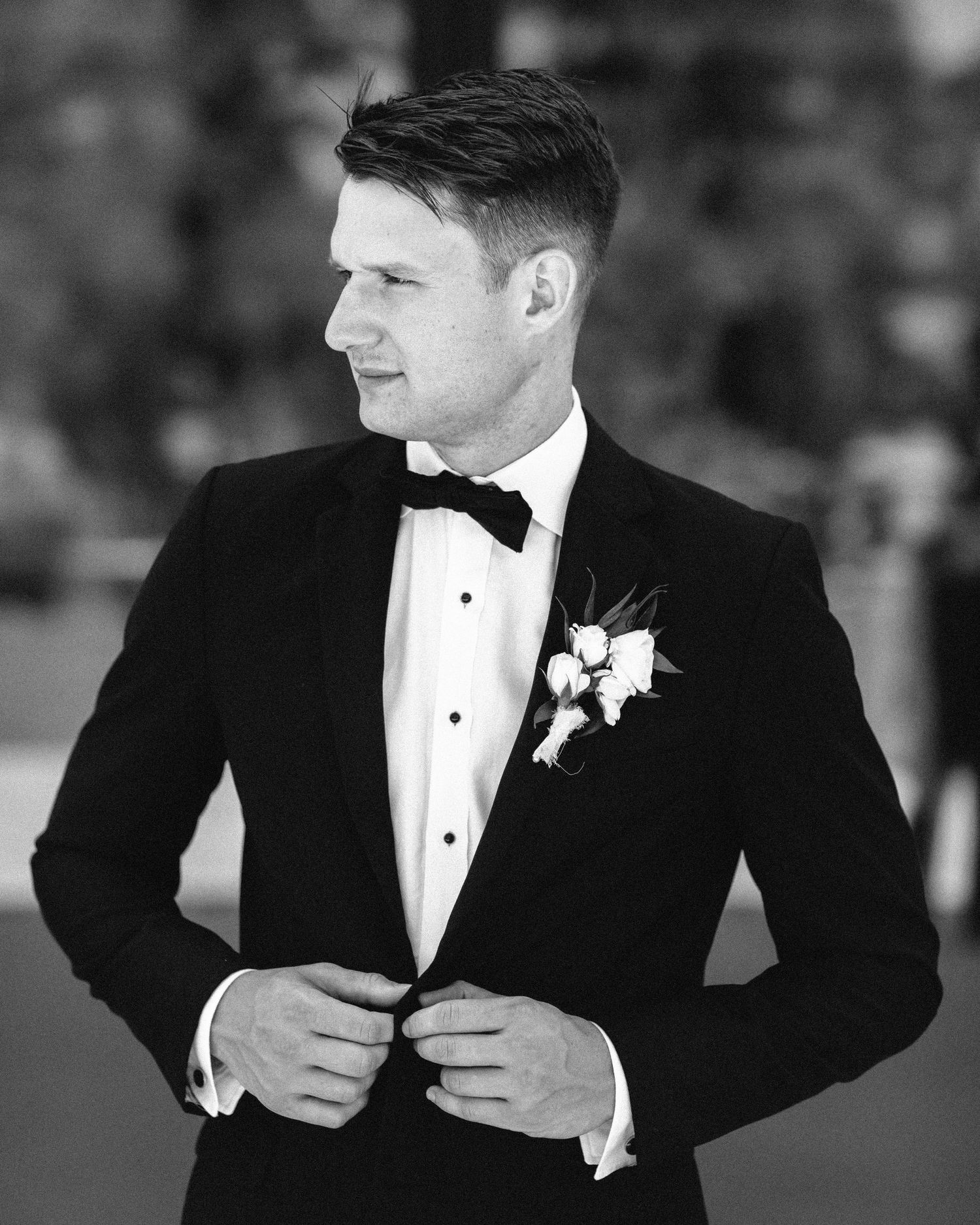 Black and white portrait of groom adjusting his tuxedo jacket with a boutonniere pinned to his lapel.