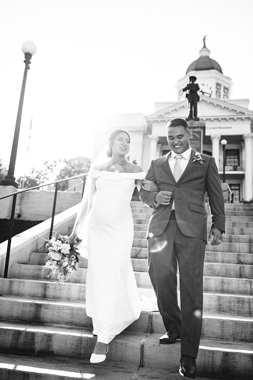 Black-and-white photo of a bride and groom walking down courthouse steps, backlit by the sun.