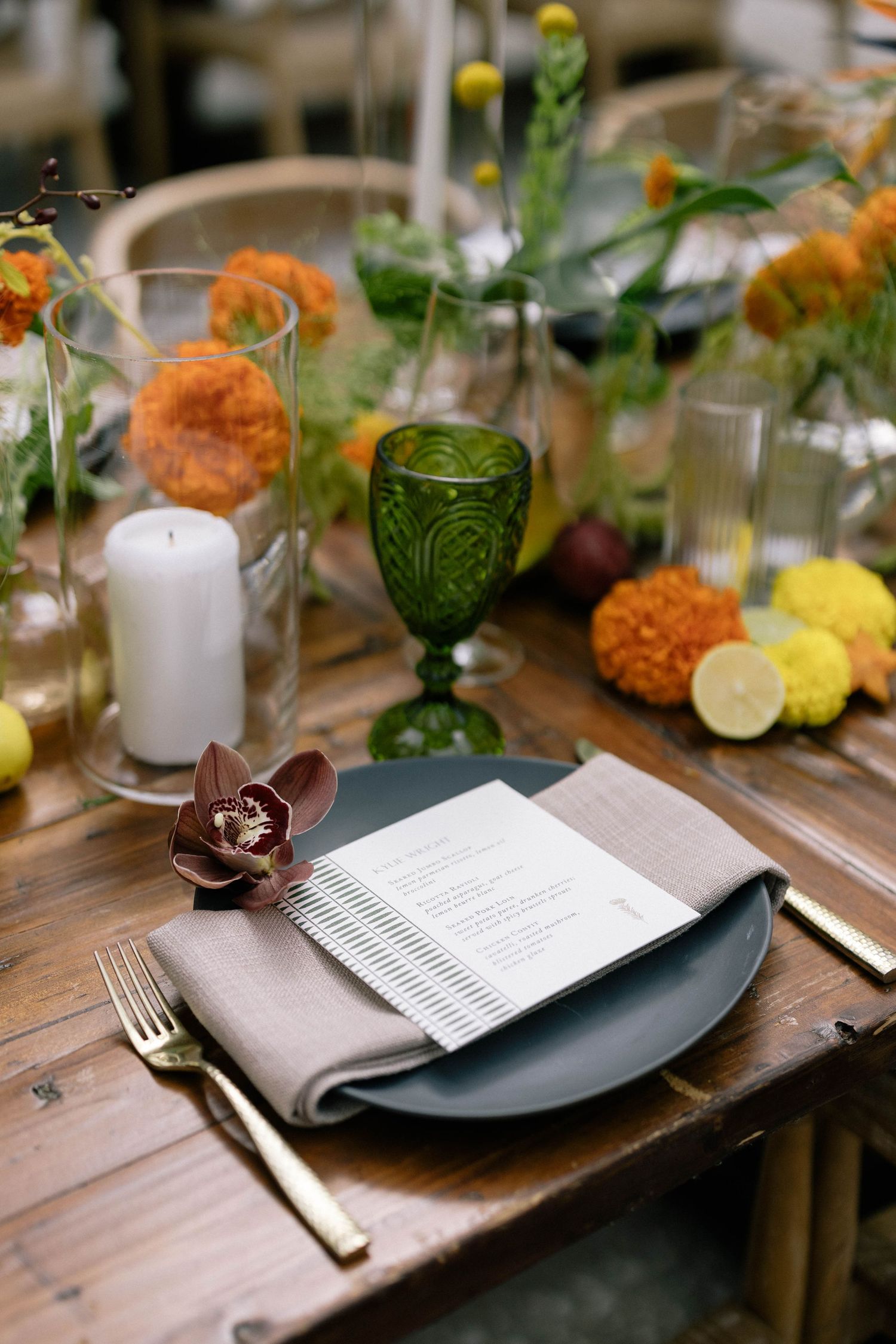 Elegant wedding table setting with a gray plate, linen napkin, and a menu topped with a burgundy orchid, gold flatware with pillar candles