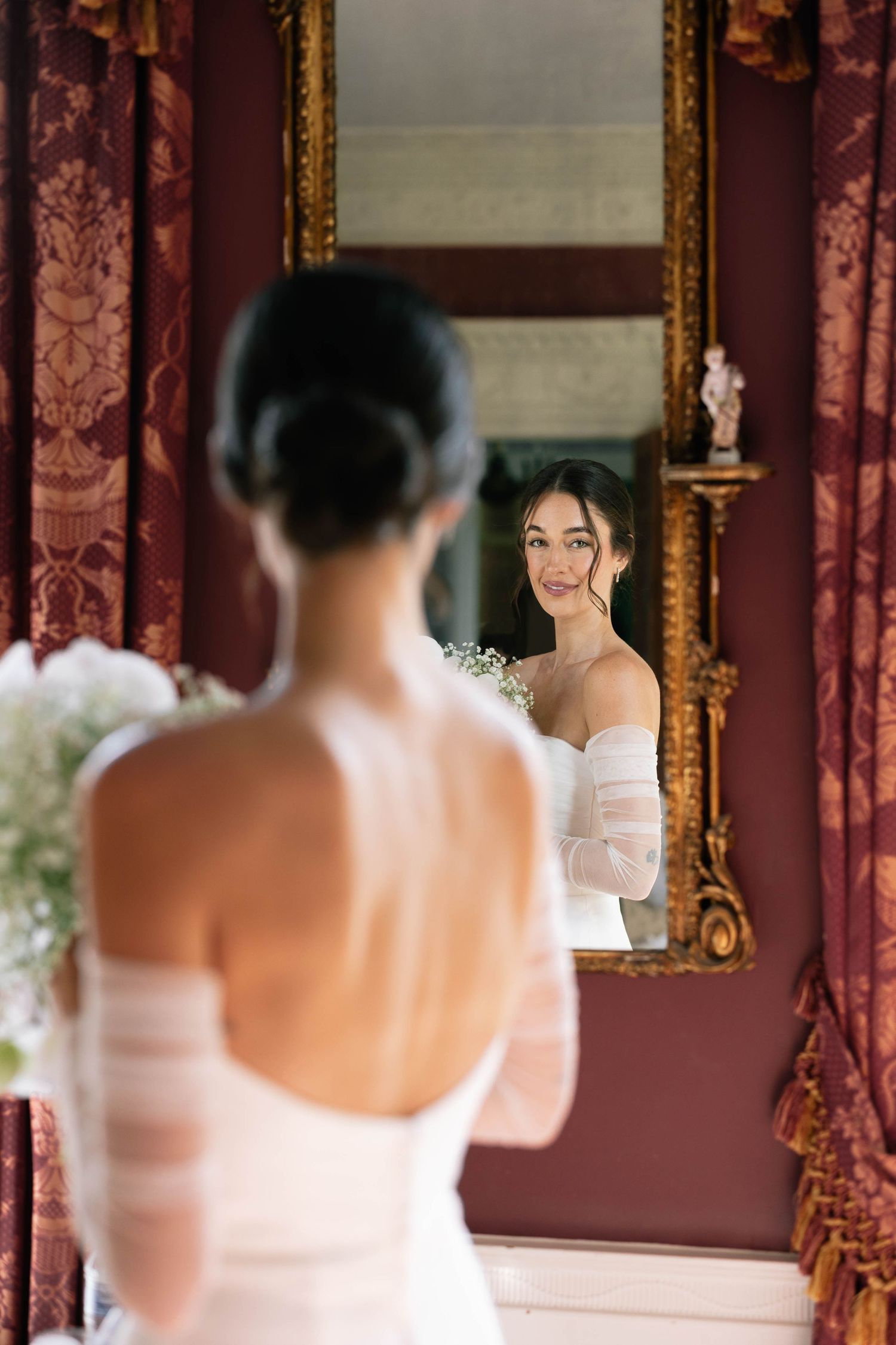 a brunette bride with an open back dress looking at herself in the mirror while holder flowers at William Aiken House, Charleston SC