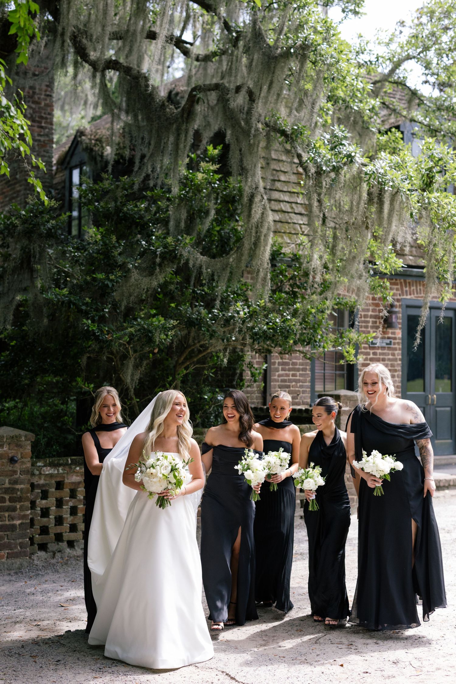 Bride in a strapless gown holding white florals, walking joyfully with her bridesmaids dressed in black gowns under moss-draped trees.