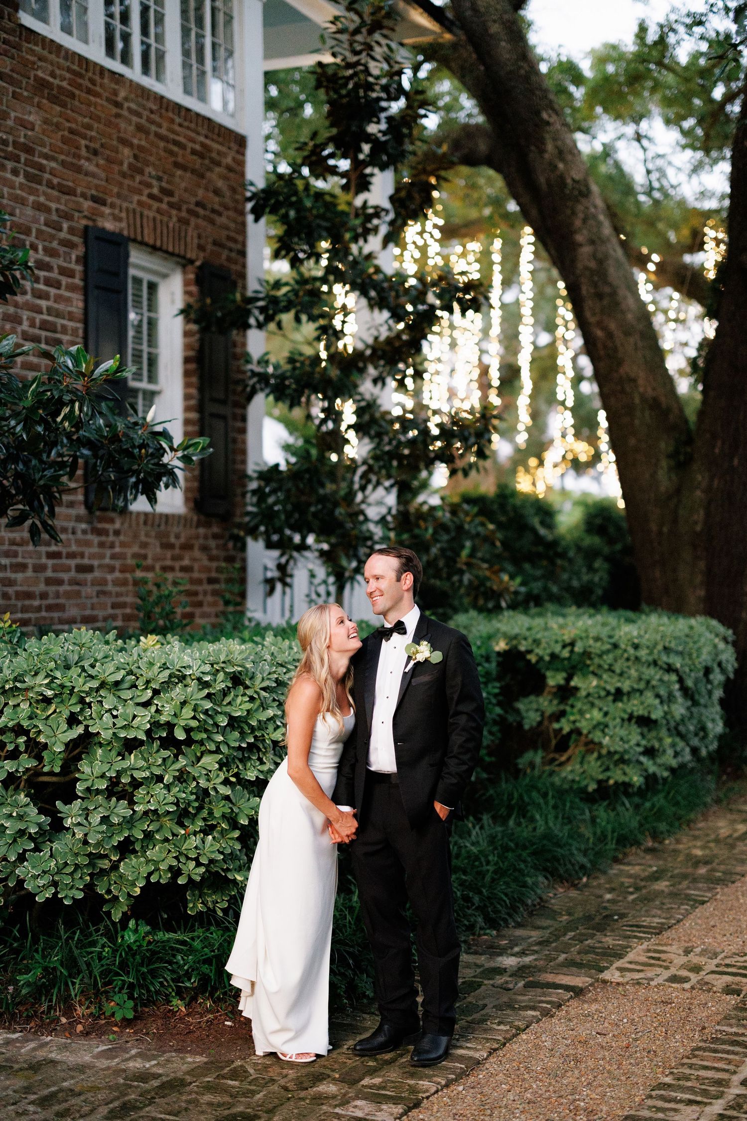 Bride in a sleek white gown and groom in a tuxedo share a lighthearted moment, holding hands in front of a brick home with twinkling string
