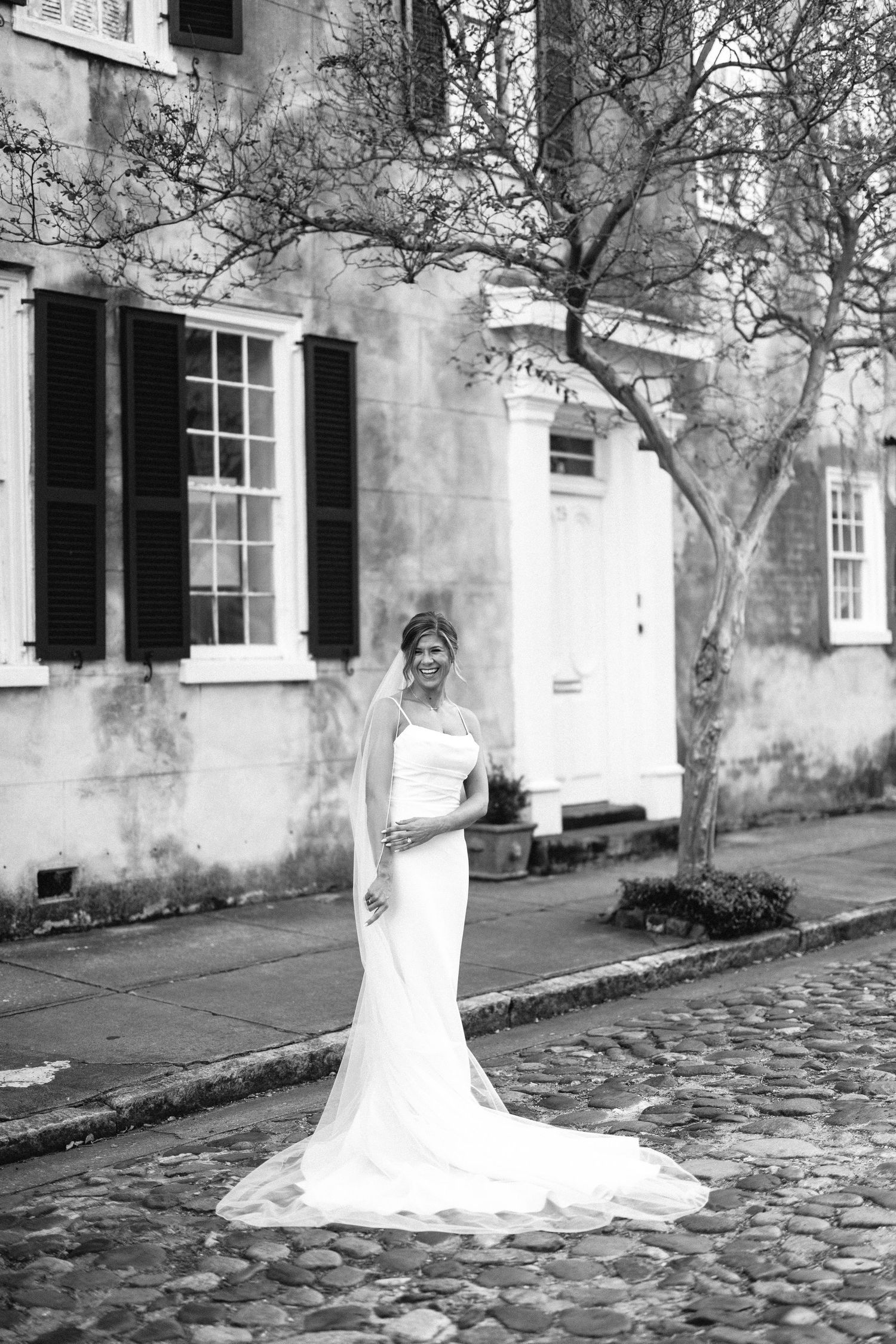 Bride in a fitted gown and veil smiles on a cobblestone street, framed by historic Charleston architecture and shutters
