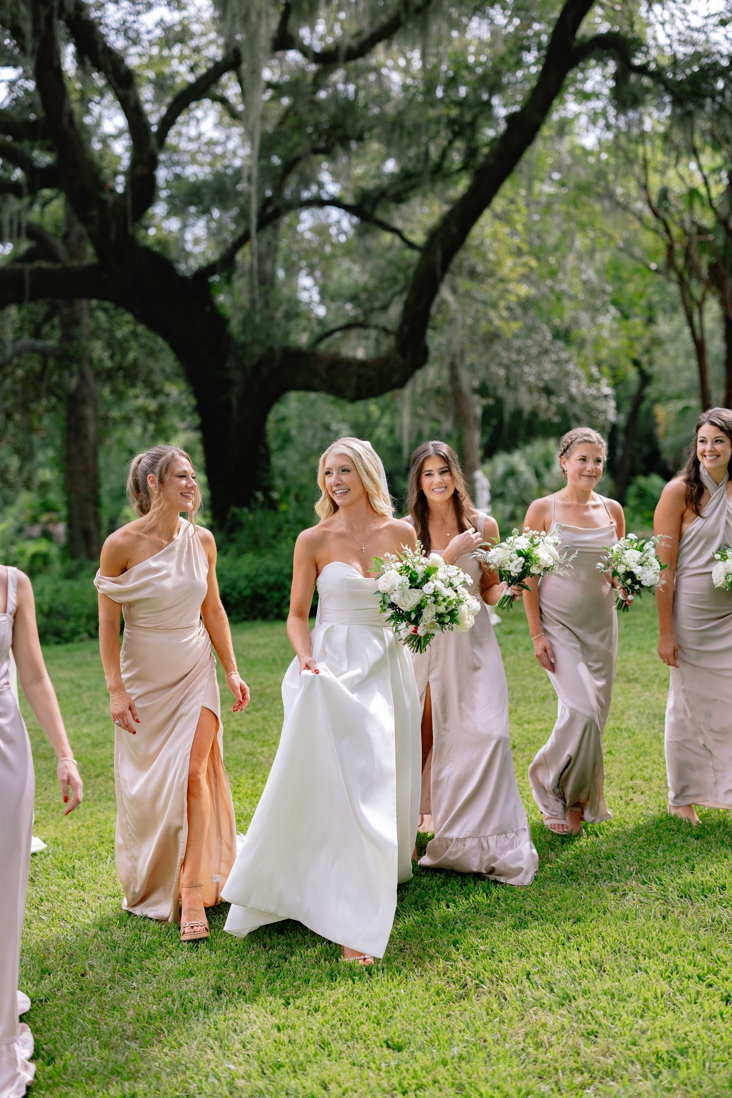 a bride smiling and walking along lush grass with her bridesmaids dressed in blush; candidly laughing in front of a southern live oak tree