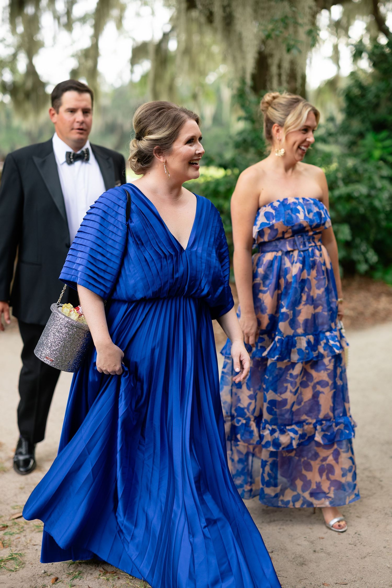 Wedding guests smiling and walking outdoors, with two women in vibrant upscale blue dresses and a man in a tuxedo.