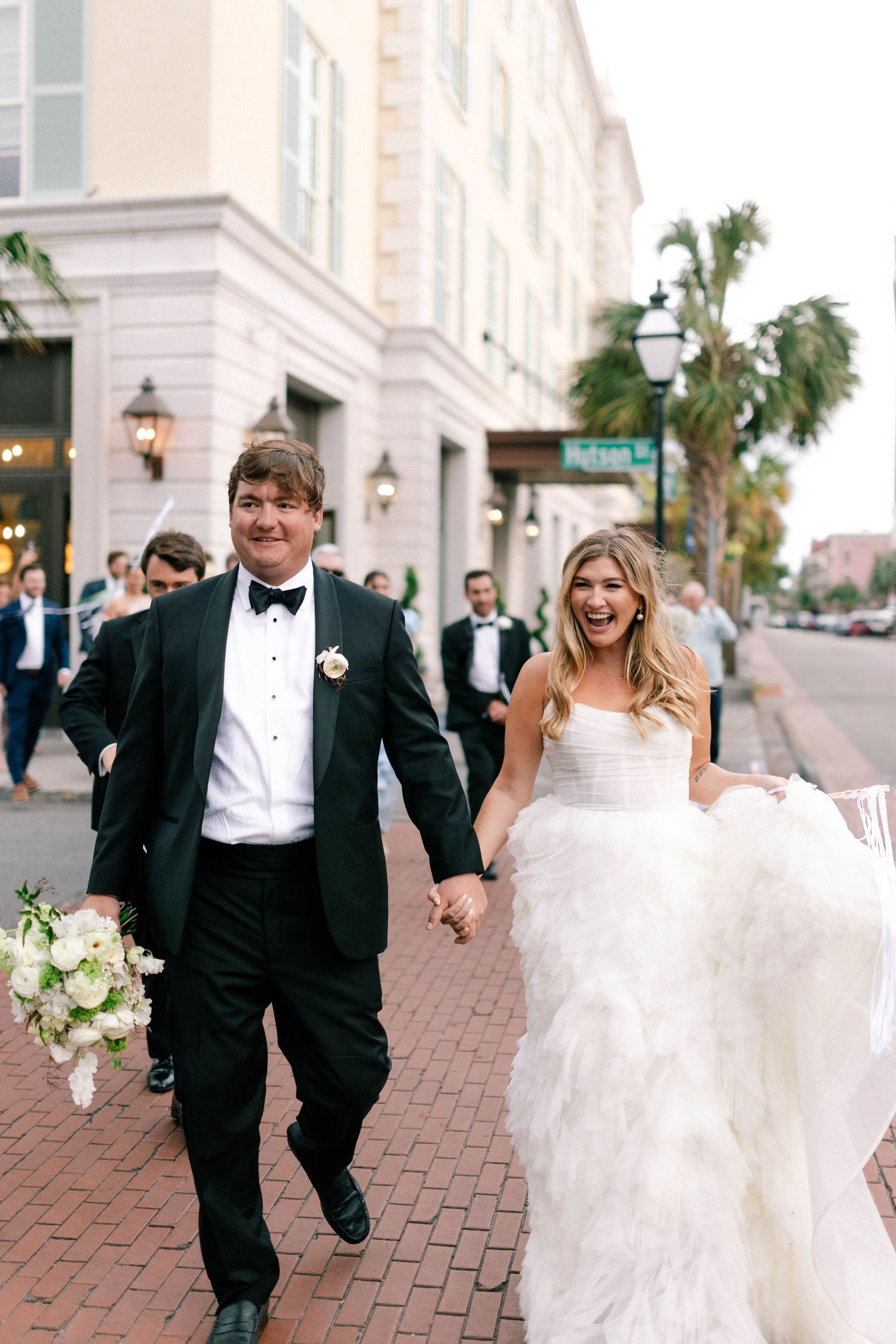 Bride and groom walking hand-in-hand down a Charleston, SC street, both laughing joyfully.