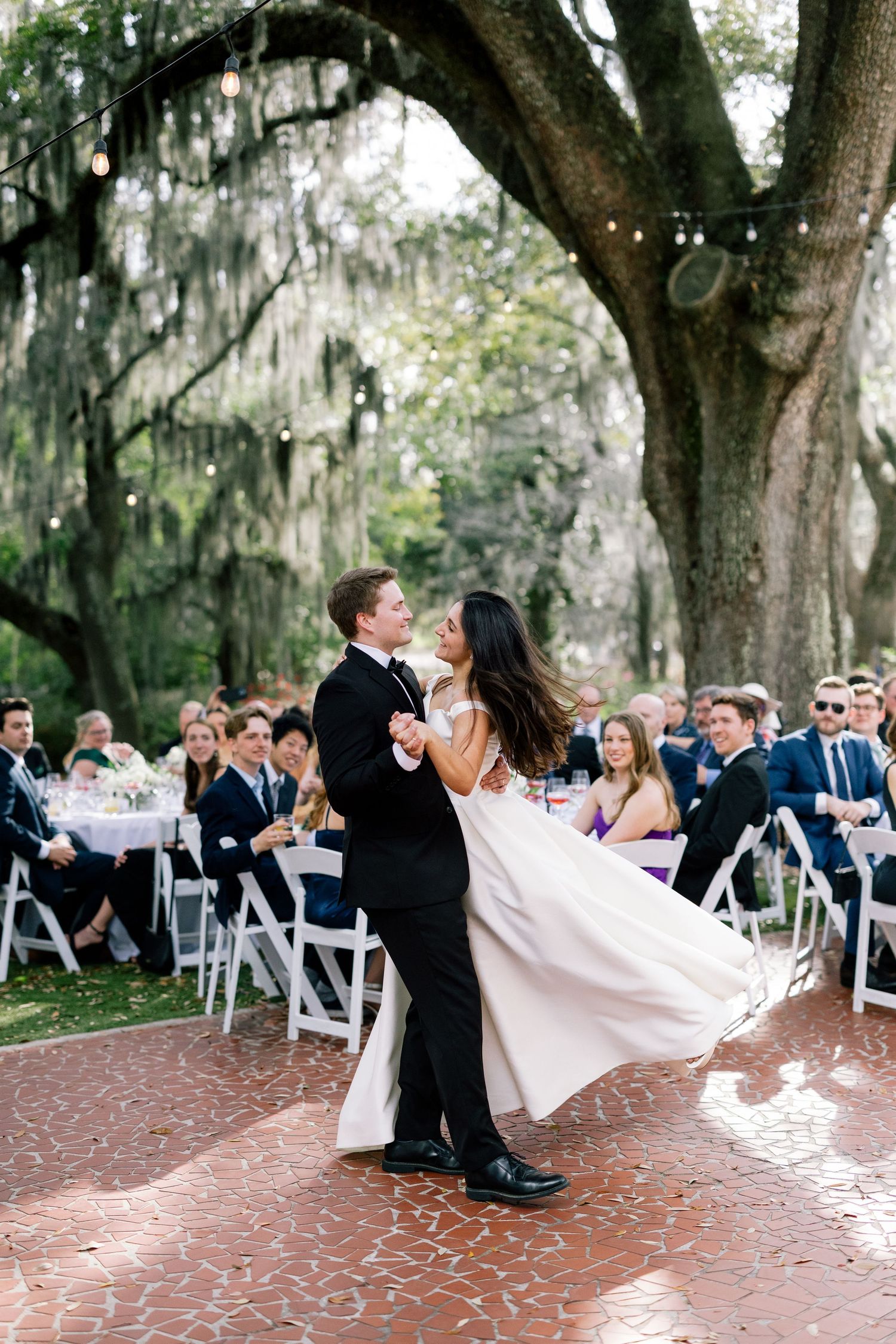 Bride and groom share a joyful first dance under oak trees with Spanish moss, surrounded by guests seated at reception tables.