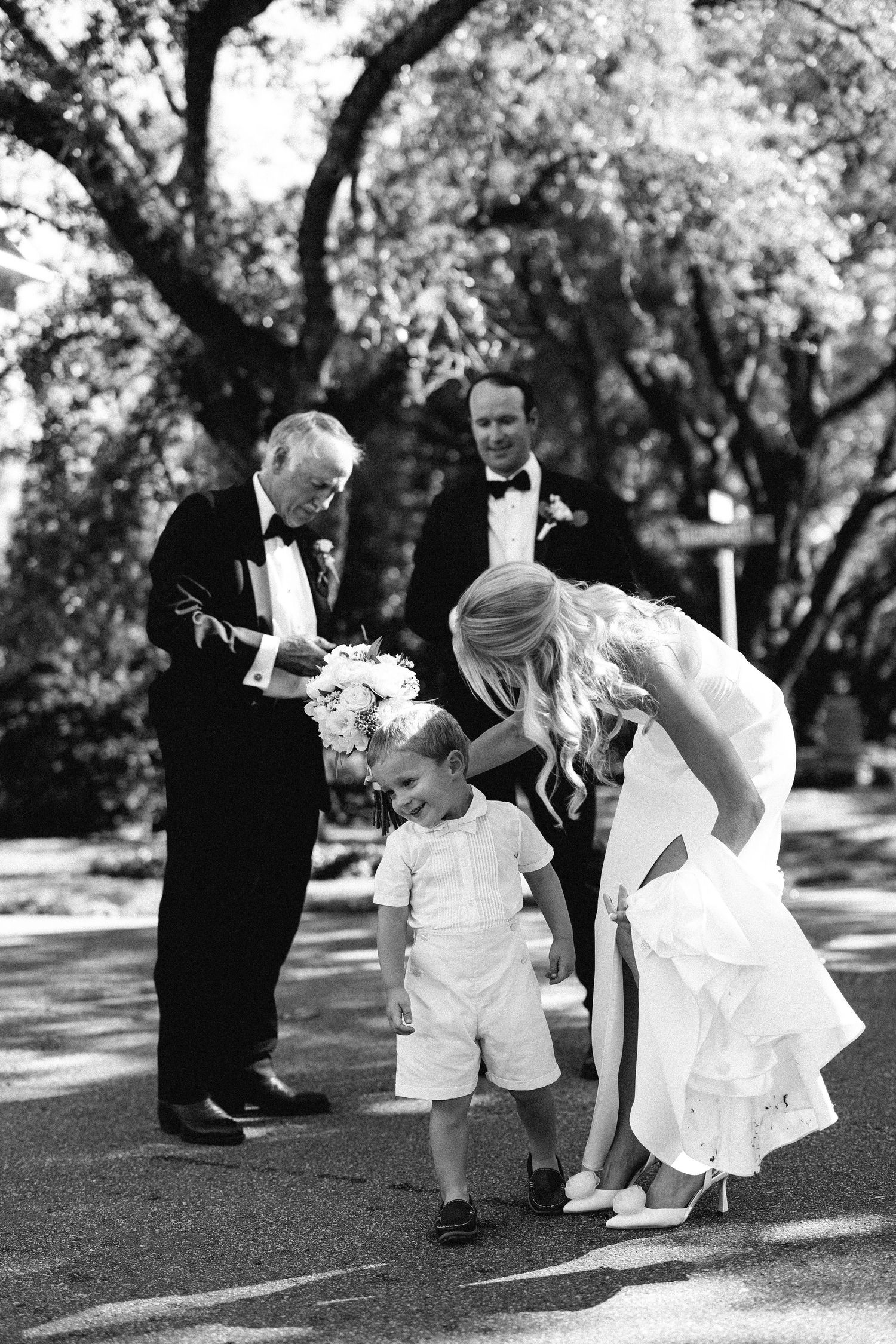 Bride in white gown bending down to smile at young ring bearer, with groomsmen in tuxedos in the background during outdoor wedding.