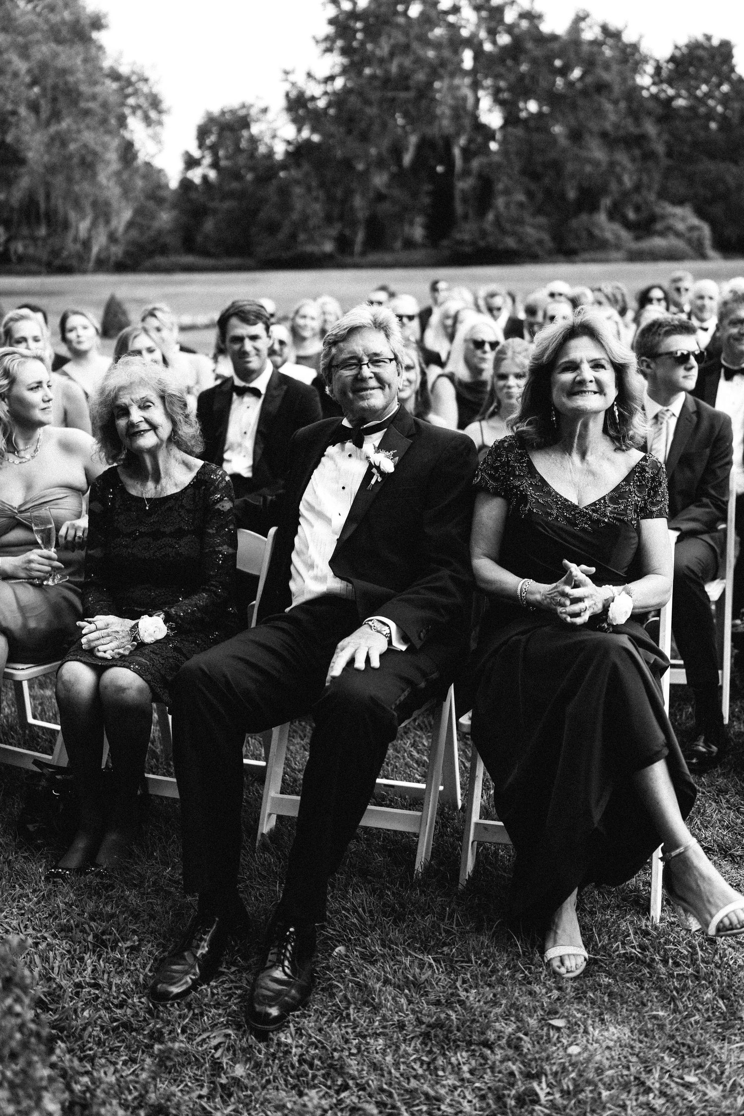 A candid black and white image of wedding guests seated outdoors, with an elderly woman, man, and woman smiling in the front row.