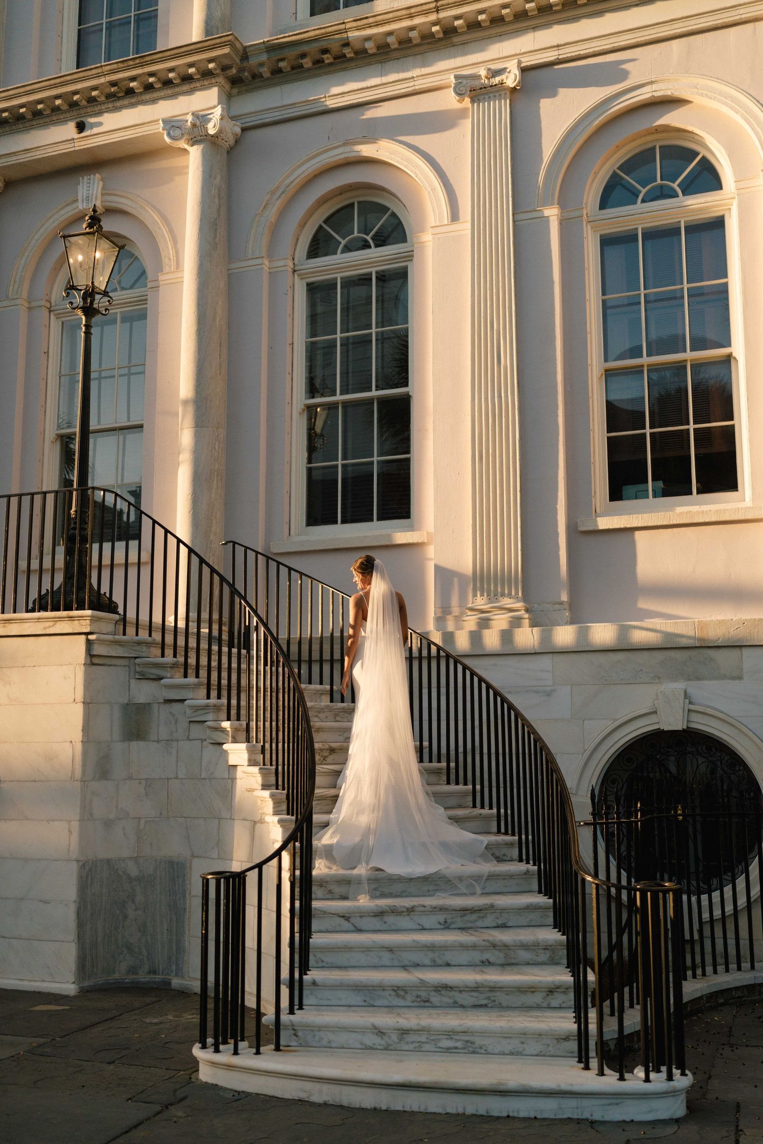 Bride in a long veil ascends a grand marble staircase of a historic Charleston building, lit by warm golden-hour sunlight