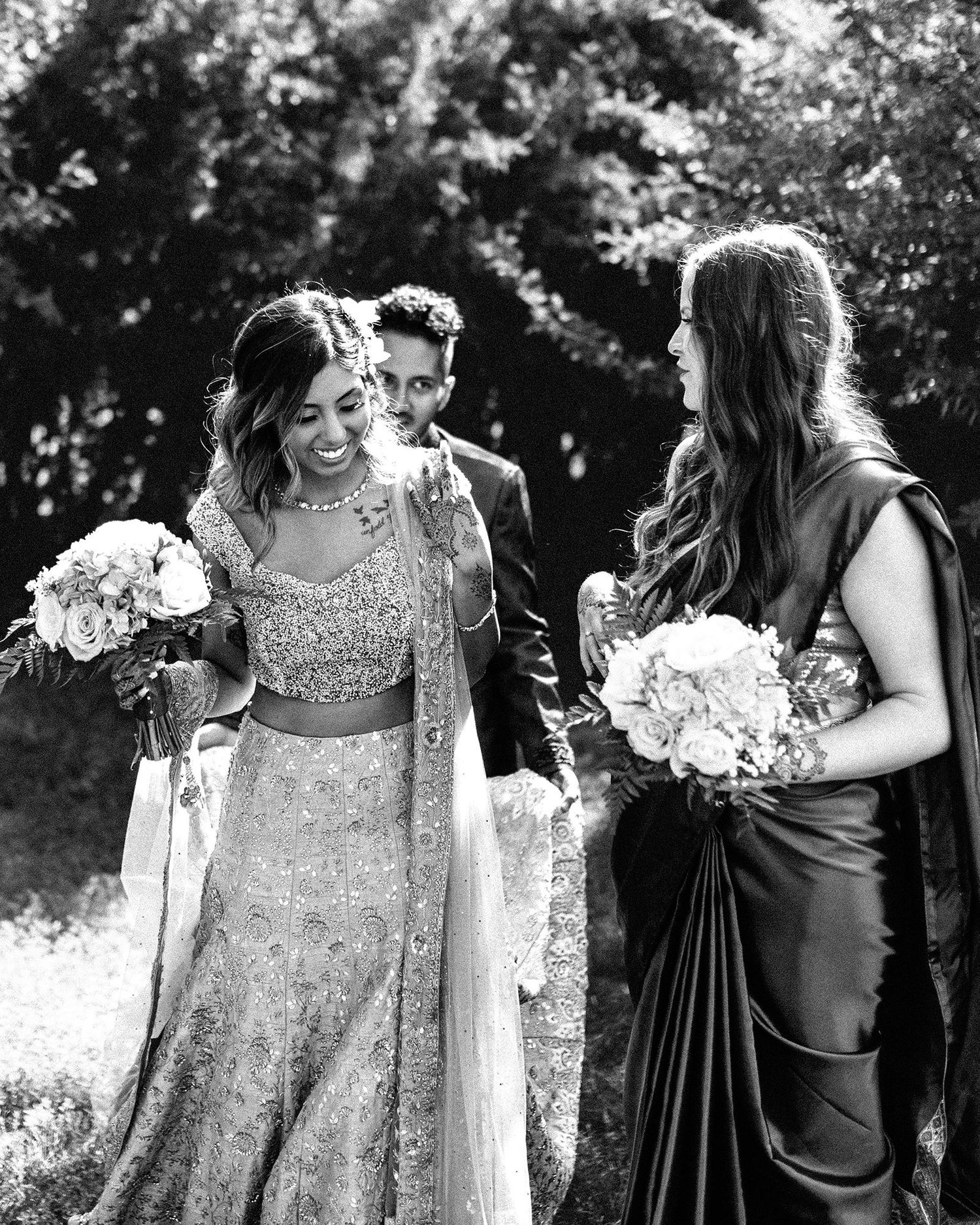Bride in an embroidered lehenga smiles while walking with bridesmaids holding bouquets during an outdoor wedding celebration