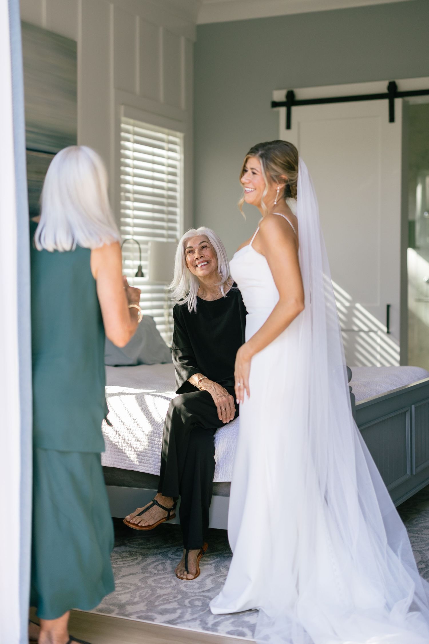 Bride in a fitted white gown with veil sharing a joyful moment with two older women in a softly lit bedroom