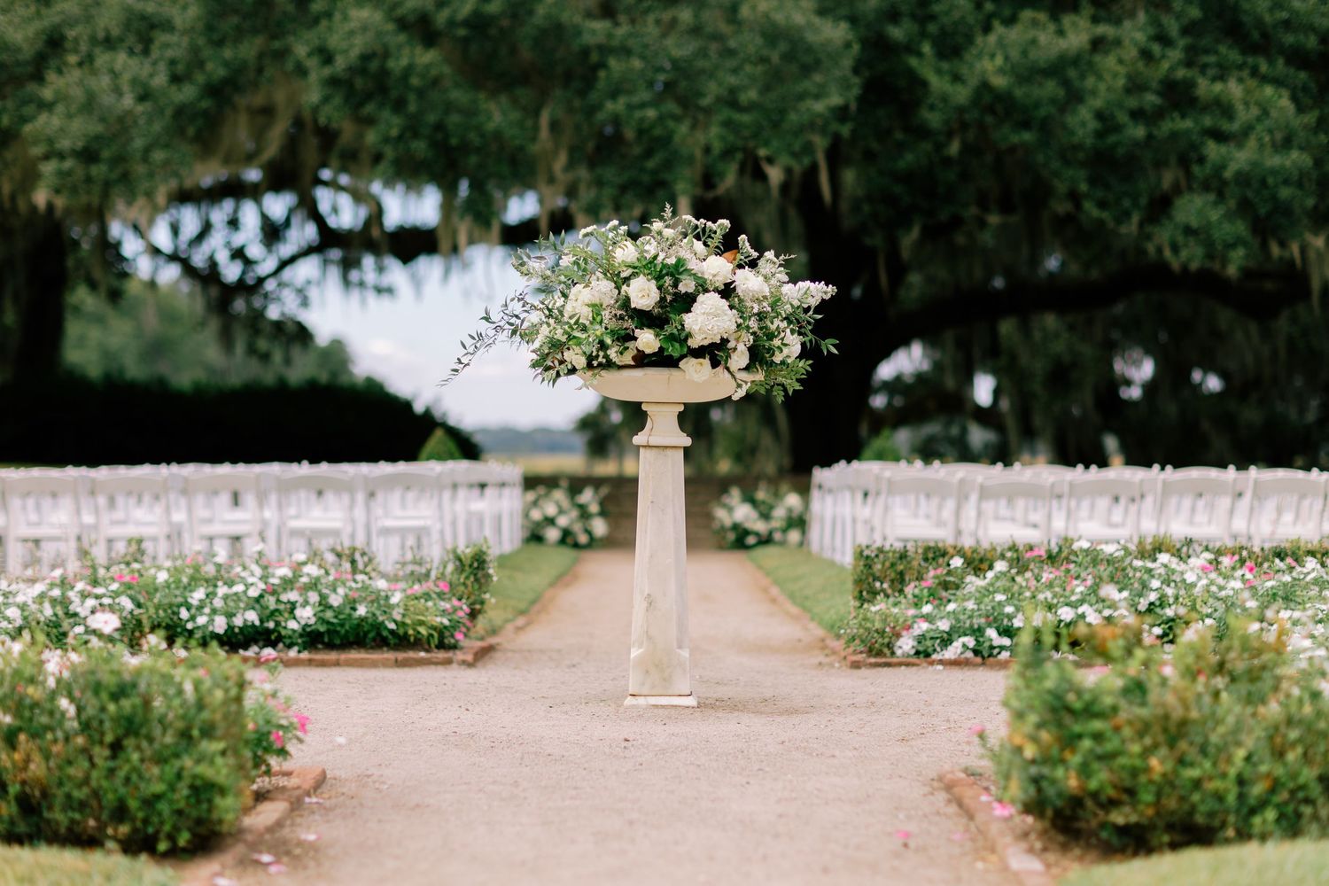 Elegant wedding ceremony setup with rows of white chairs and a tall floral arrangement on a pedestal, framed by oak trees at Middleton Place