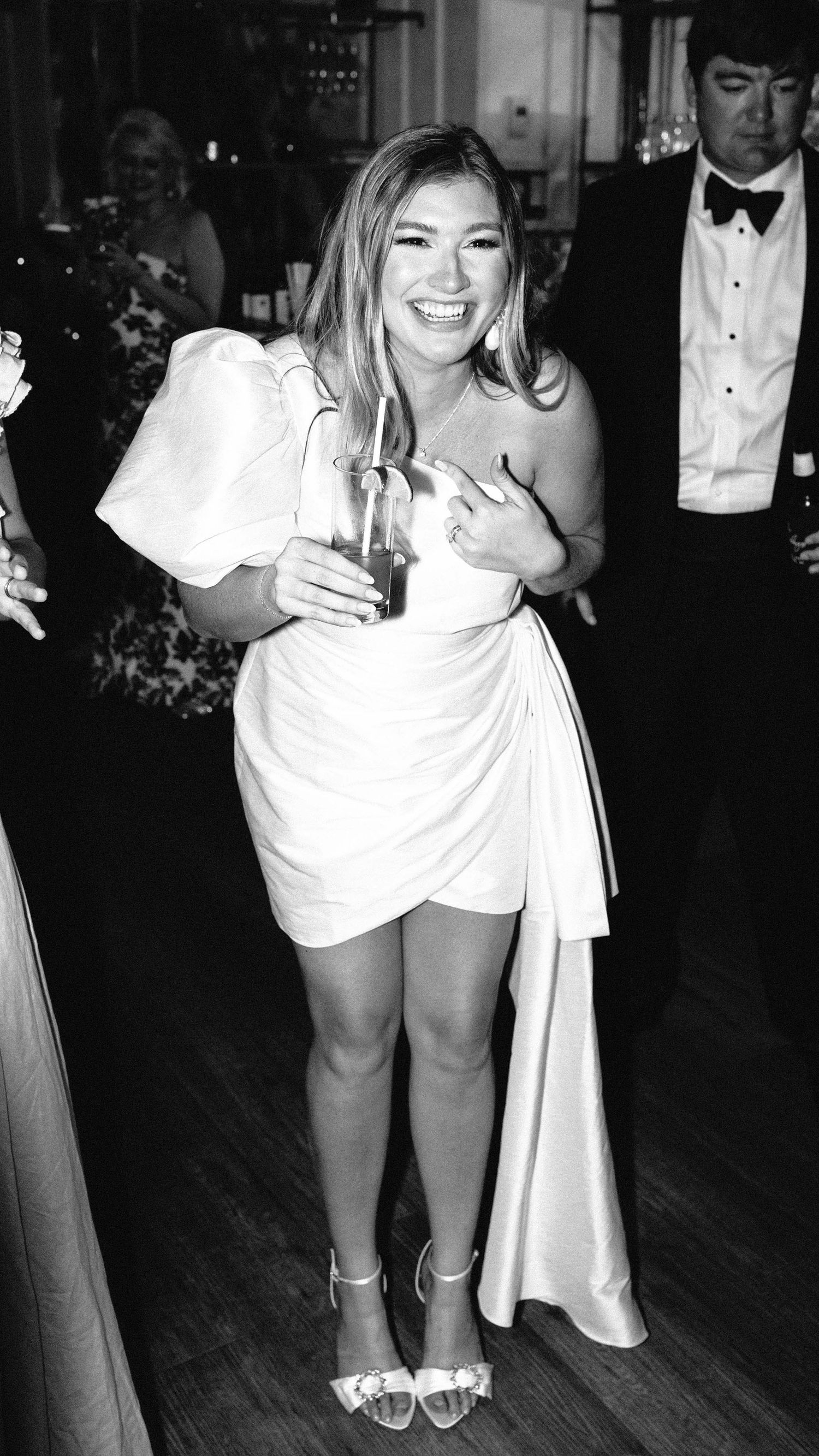 Black and white candid of a joyful bride in a short, one-shoulder dress, holding a drink and laughing on the dance floor with formal guests