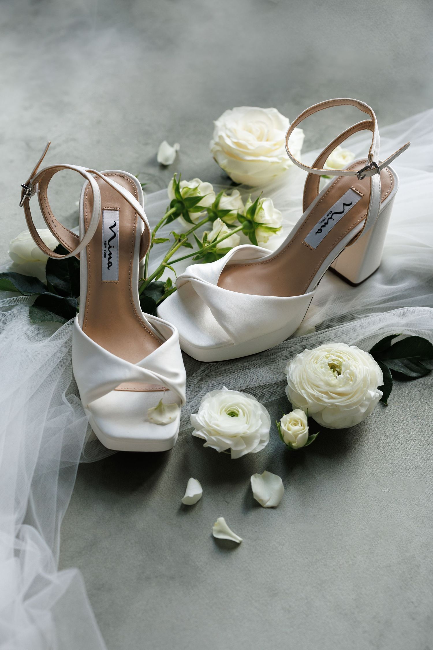 Pair of white block-heeled bridal shoes displayed with white roses and soft veil fabric for detail styling.