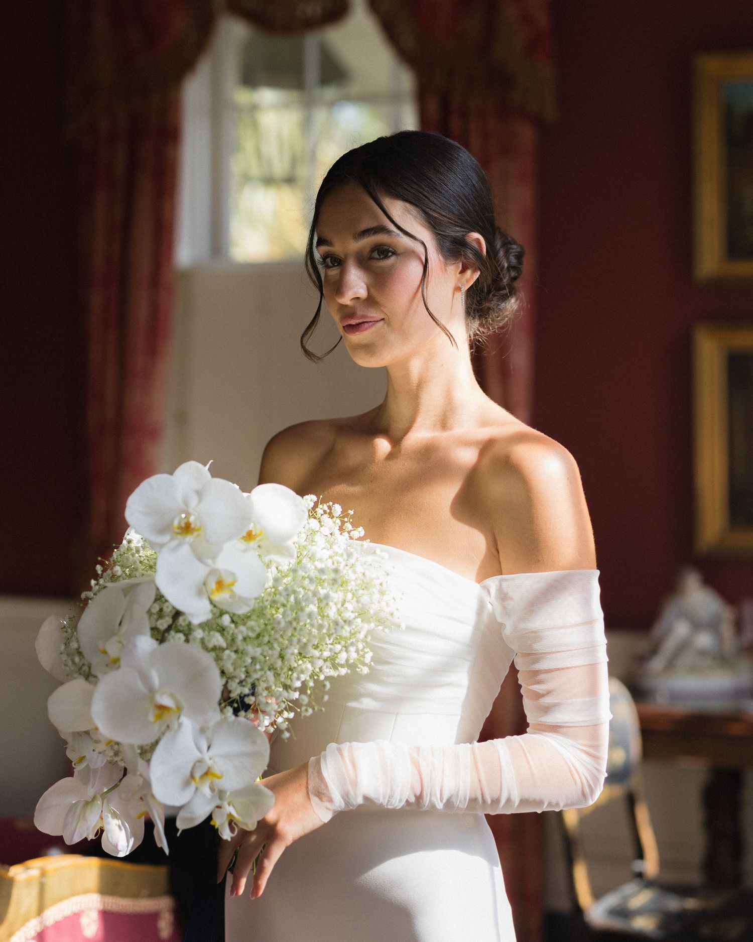 Bride in an off-the-shoulder gown holding a cascading bouquet of white orchids and baby's breath, softly lit inside William Aiken House.
