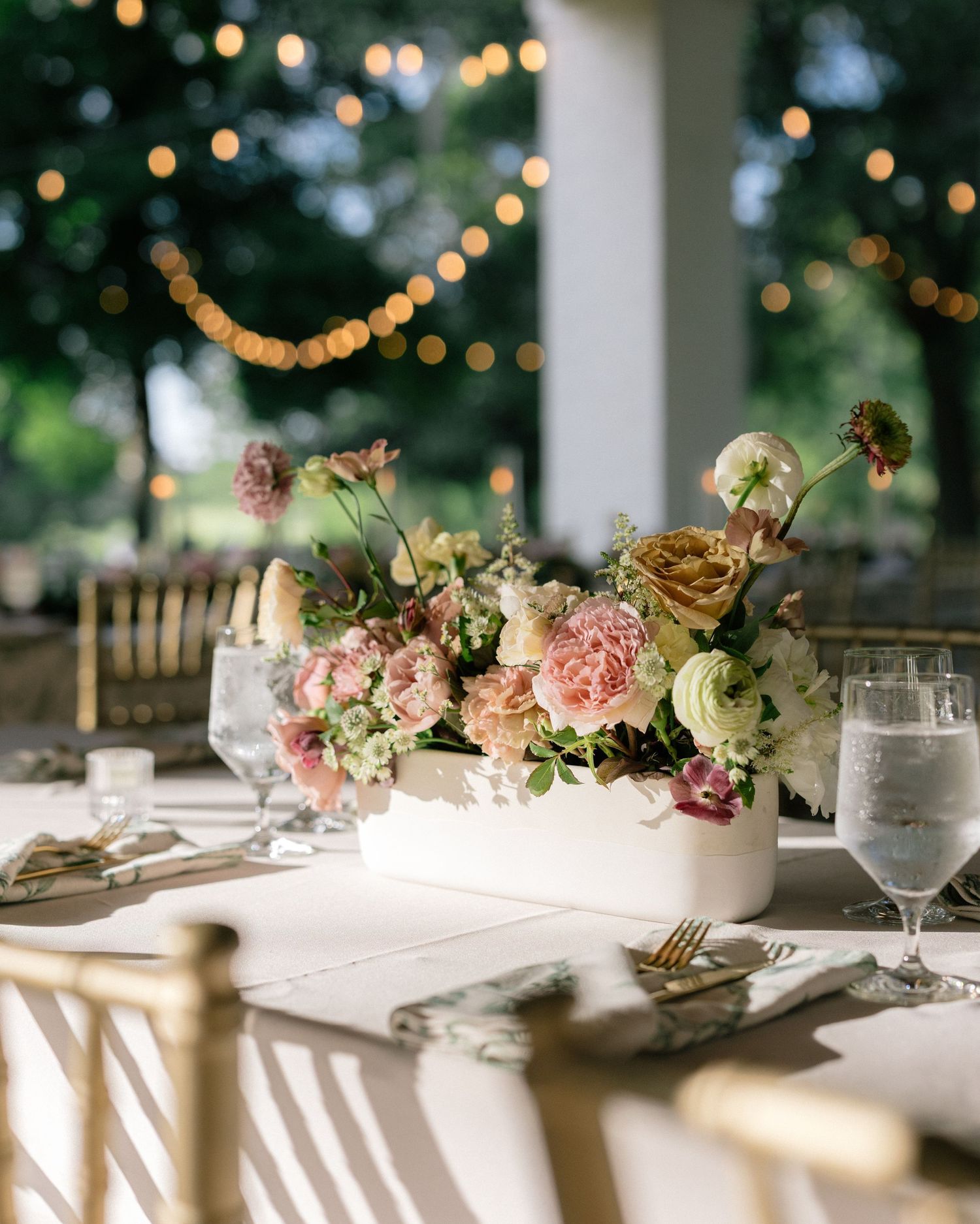 Elegant wedding reception table centerpiece with pastel flowers including roses and ranunculus in a white vase, with string lights