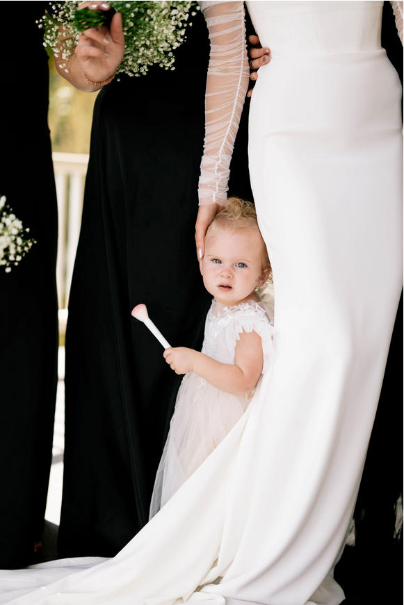 A toddler in a tulle dress peeks out while holding the bride’s gown, standing among women in black dresses holding bouquets