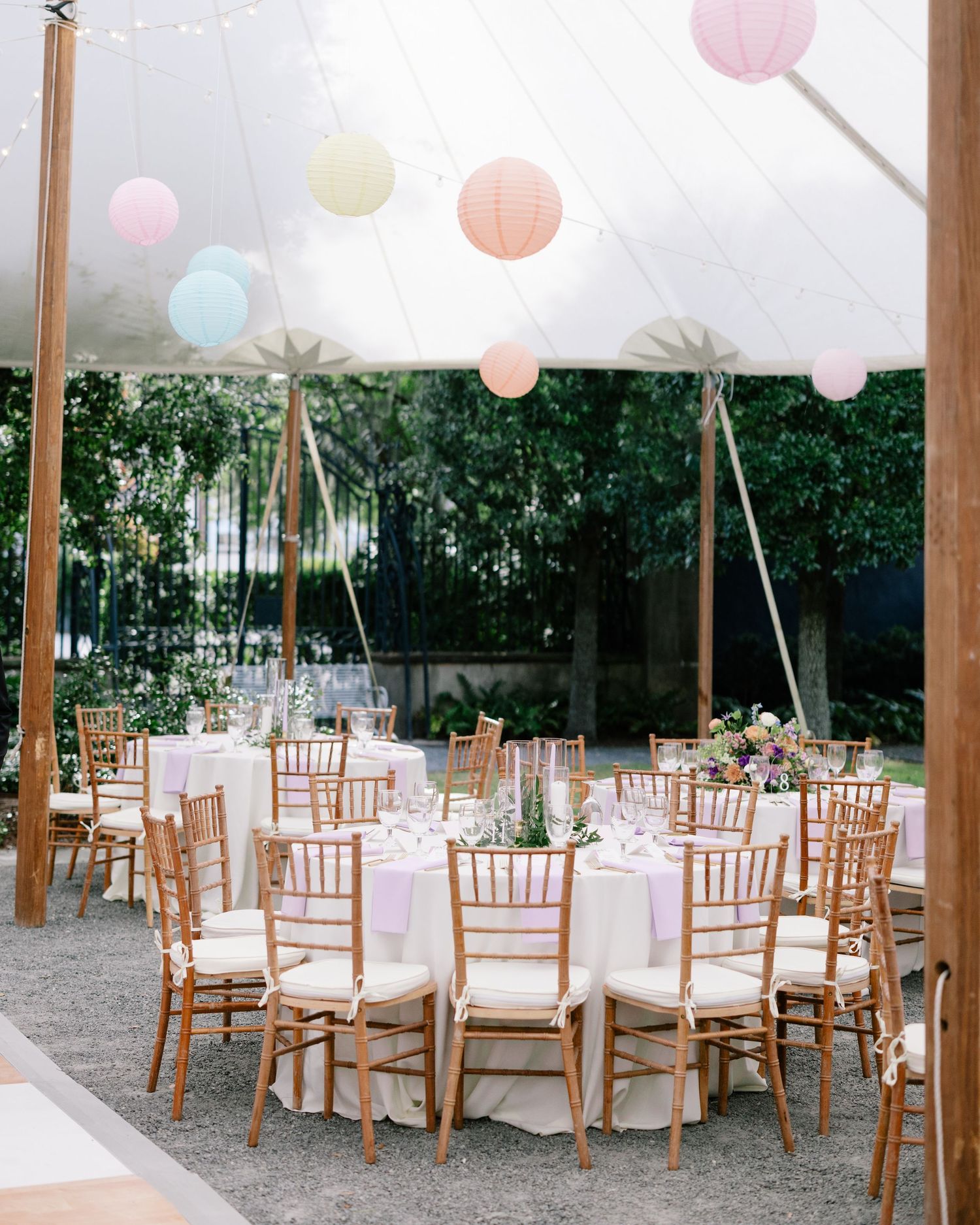 An elegant outdoor wedding reception setup under a tent with pastel paper lanterns, round tables, and wooden chairs