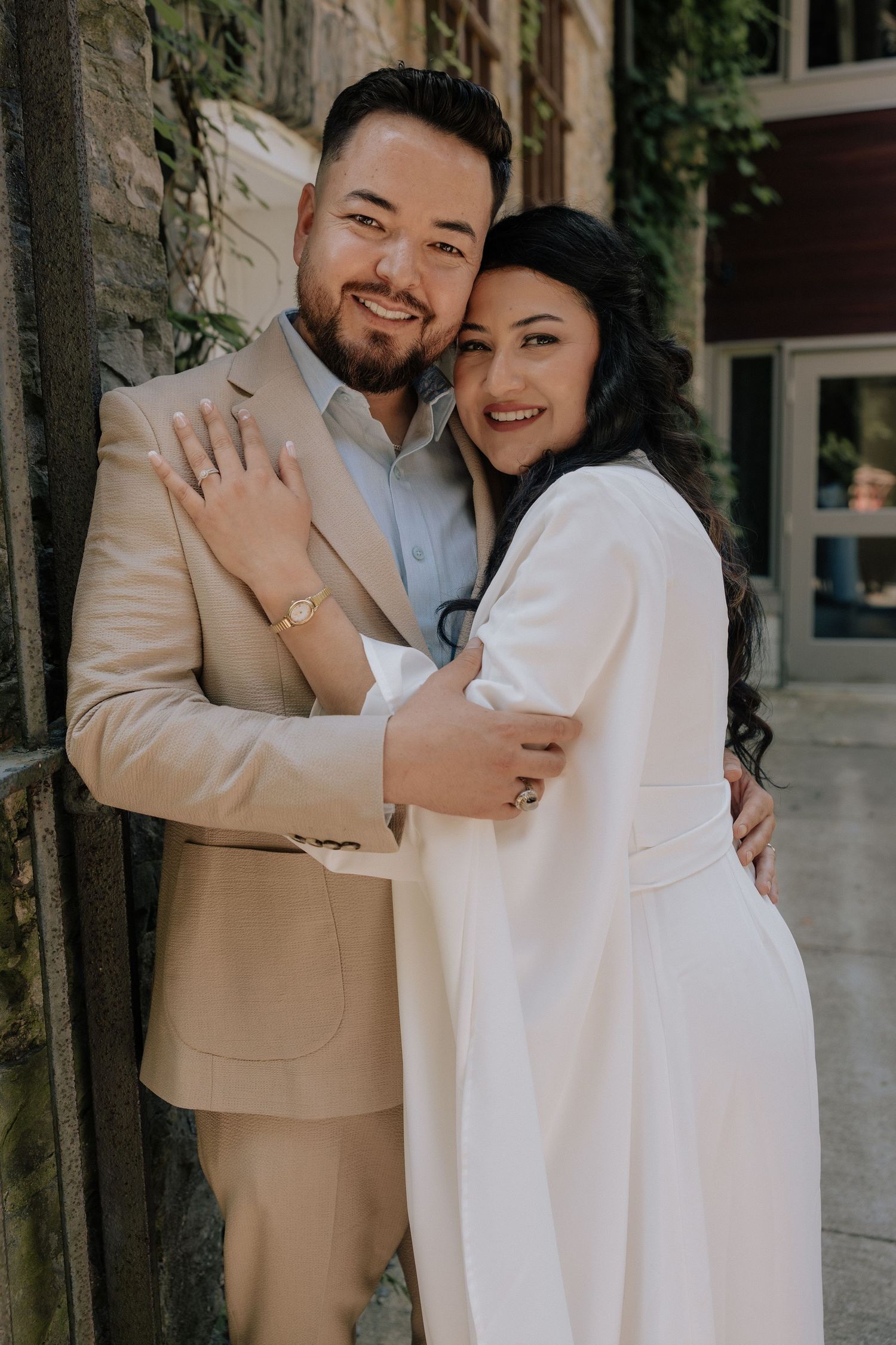 Happy engaged couple posing together, woman in white dress showing engagement ring, man in beige suit, urban backdrop.