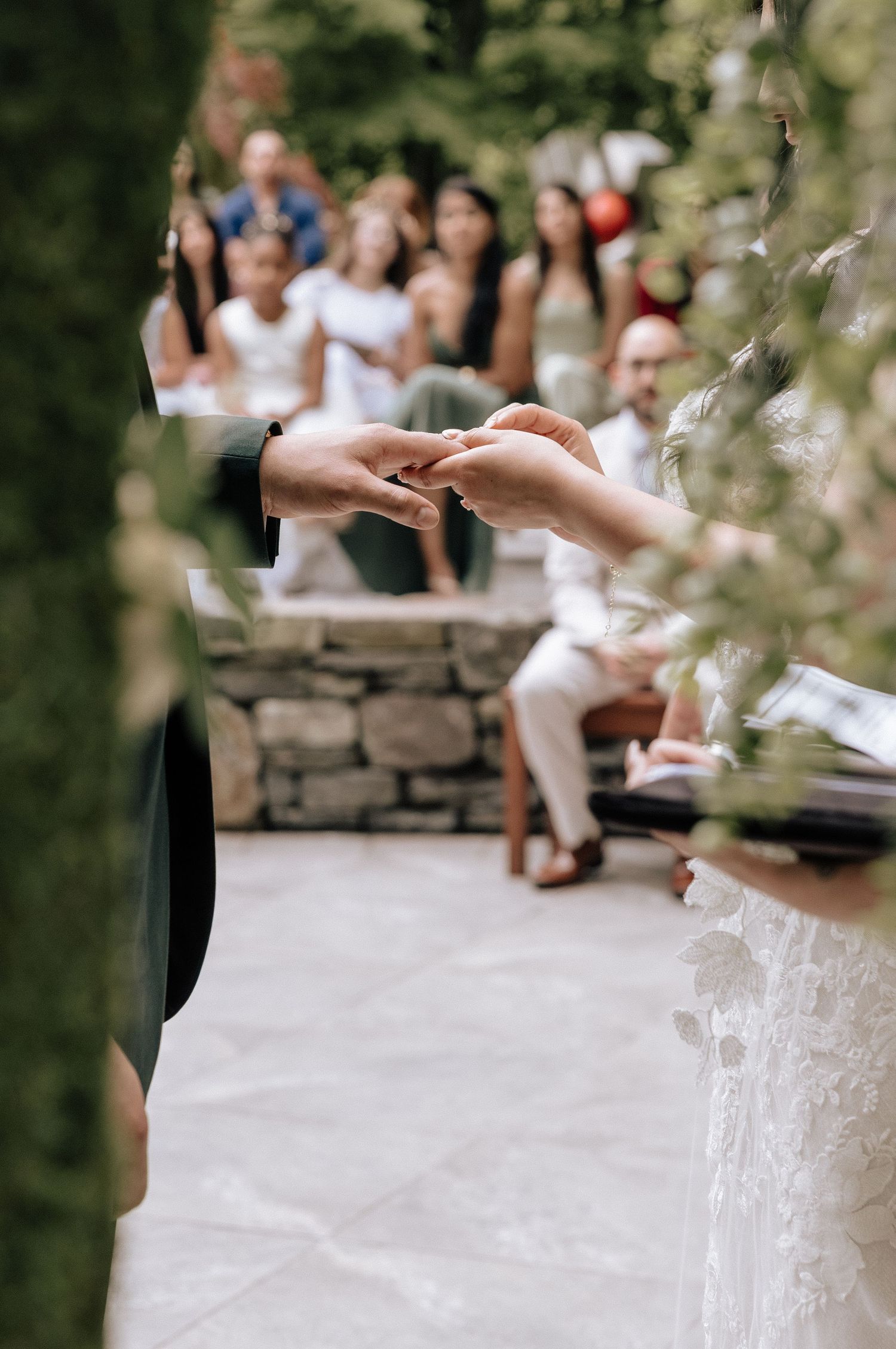 Close-up view of hands exchanging wedding rings during an outdoor ceremony with guests in background.