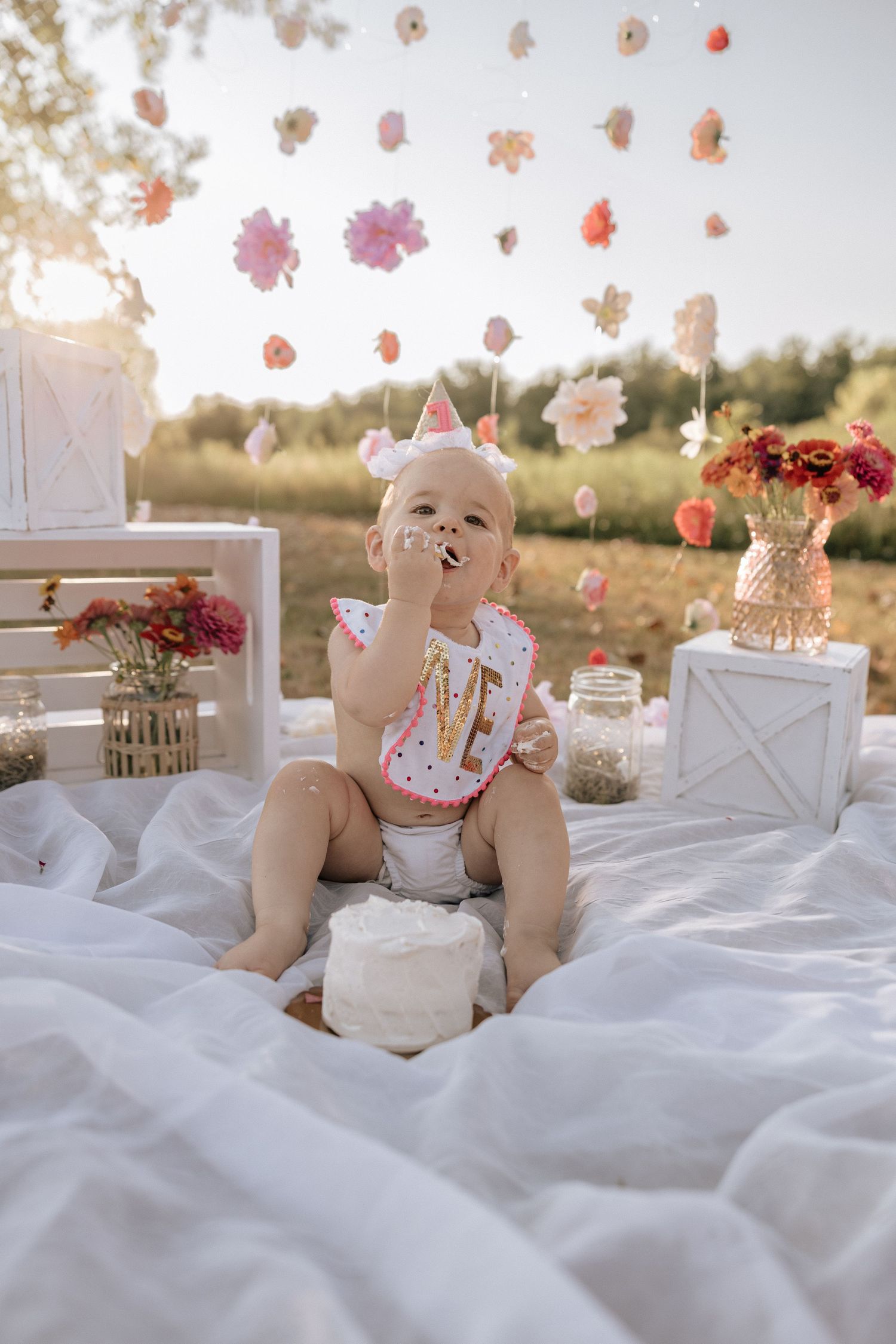 A teddy bear sits on white bedding with delicate pink floral petals falling in a dreamy outdoor setting.