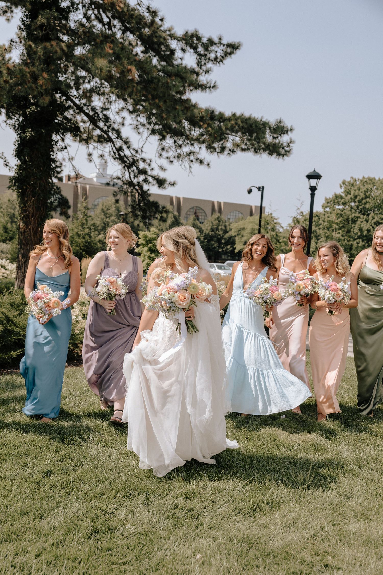 A group of bridesmaids in pastel dresses hold up their skirts while posing together on a grassy hillside.