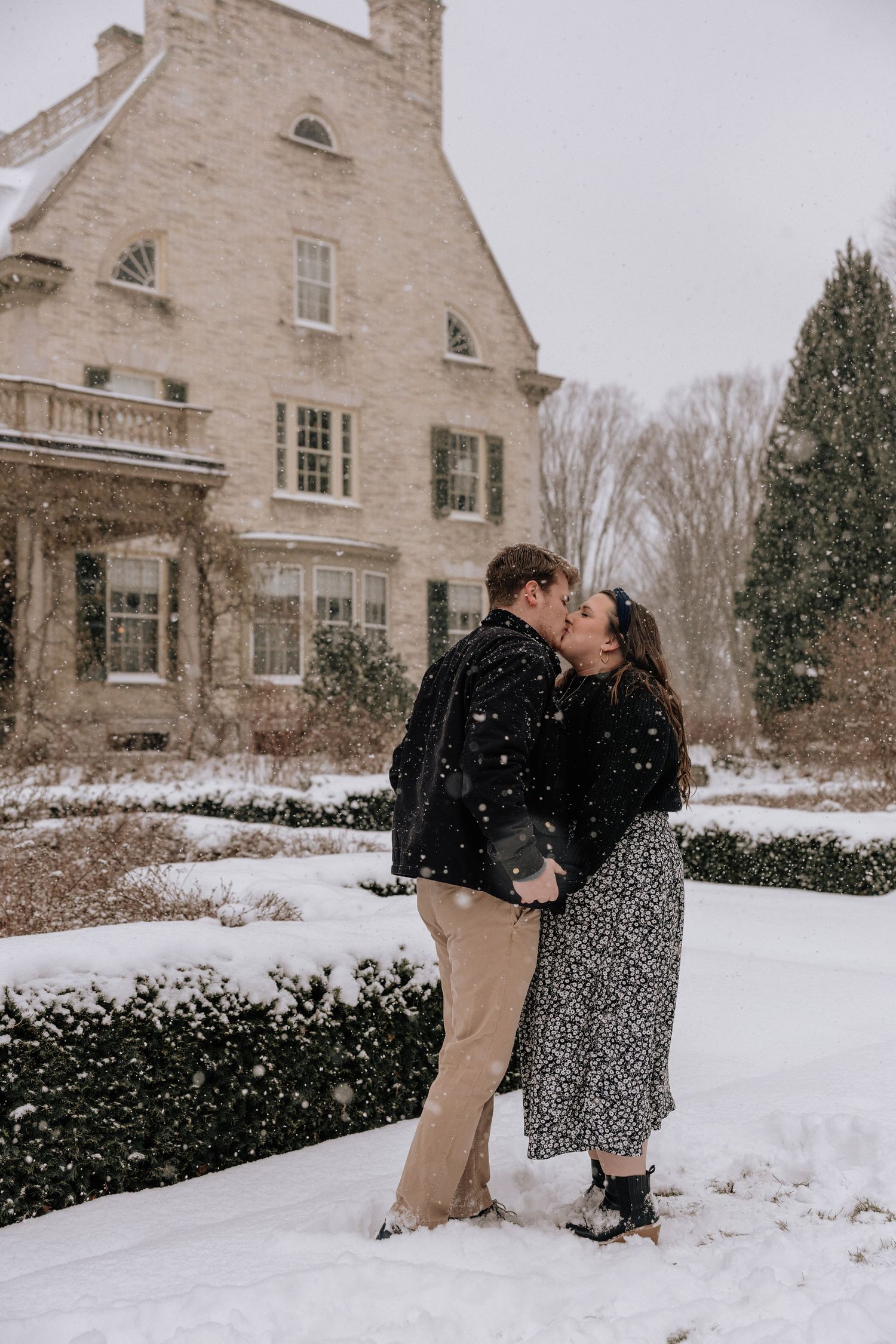 A romantic winter scene unfolds as a couple shares a kiss in front of a snow-covered stone house.