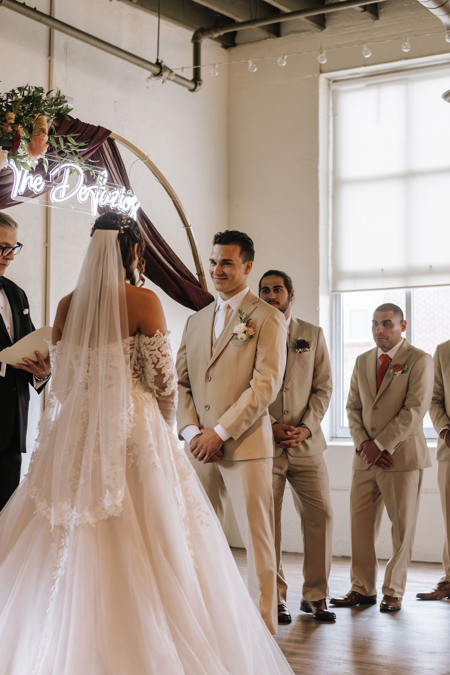 Groomsmen in tan suits gather in a bright indoor venue as a bride in a flowing white gown with veil approaches for photos.