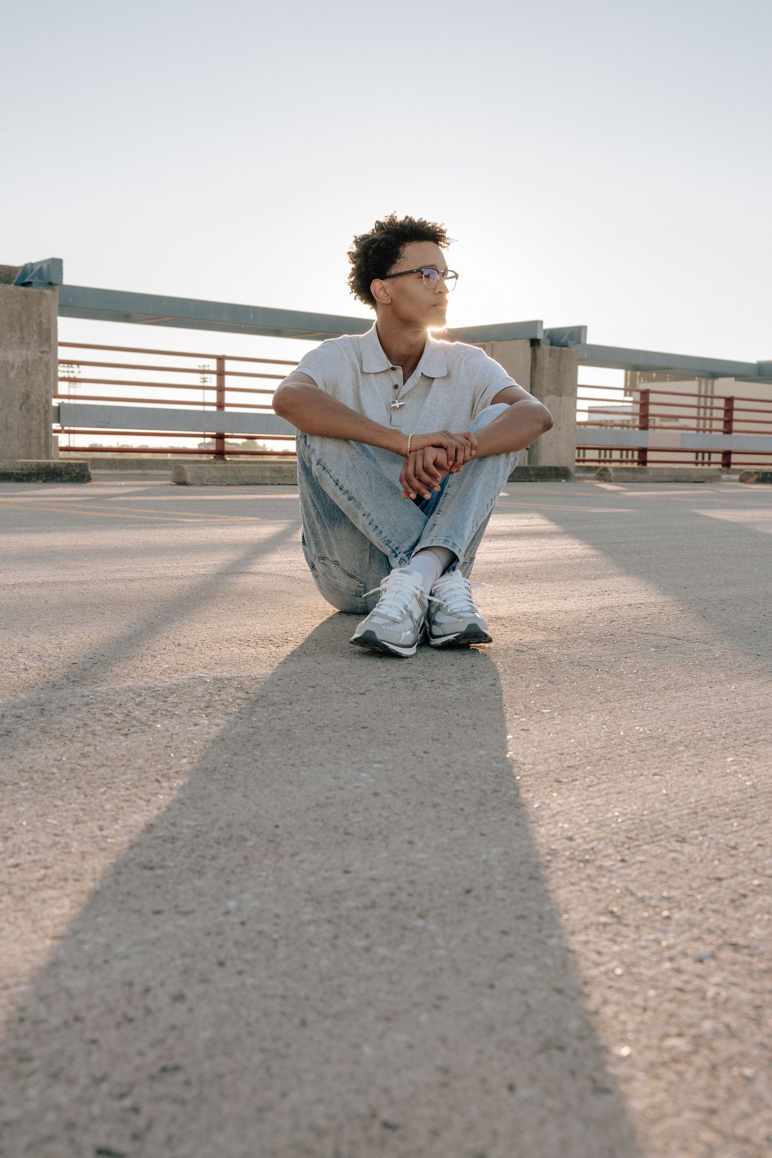 Young person with curly hair and glasses sitting on concrete ground outdoors, wearing jeans and white sneakers.