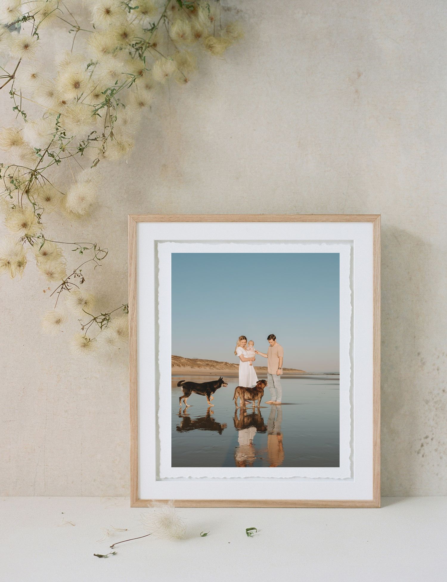 Beach photograph in wooden frame showing reflections on wet sand with delicate flowers in corner.