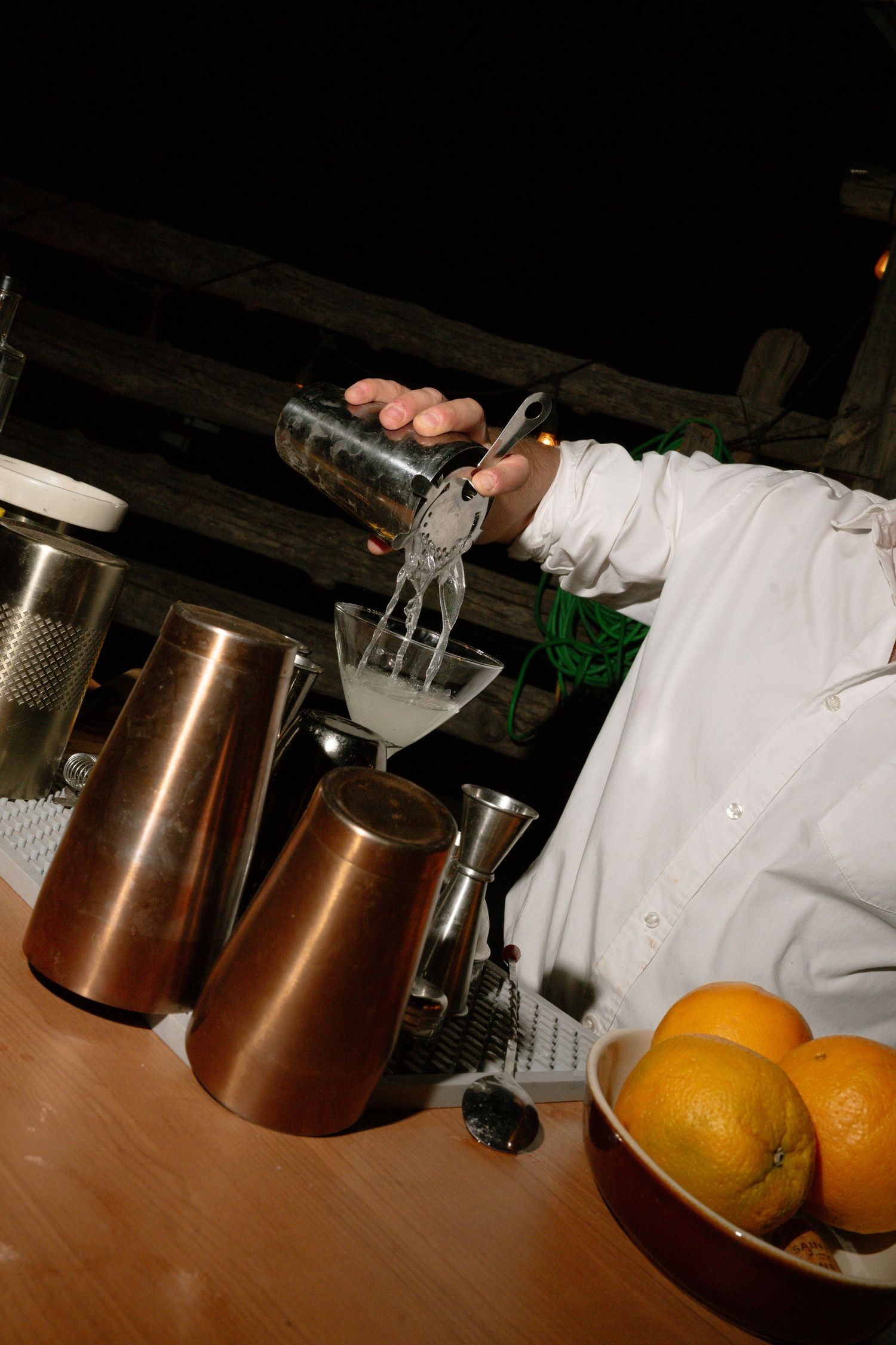 Bartender pouring cocktail shaker at outdoor wedding bar with copper vessels and fresh oranges.