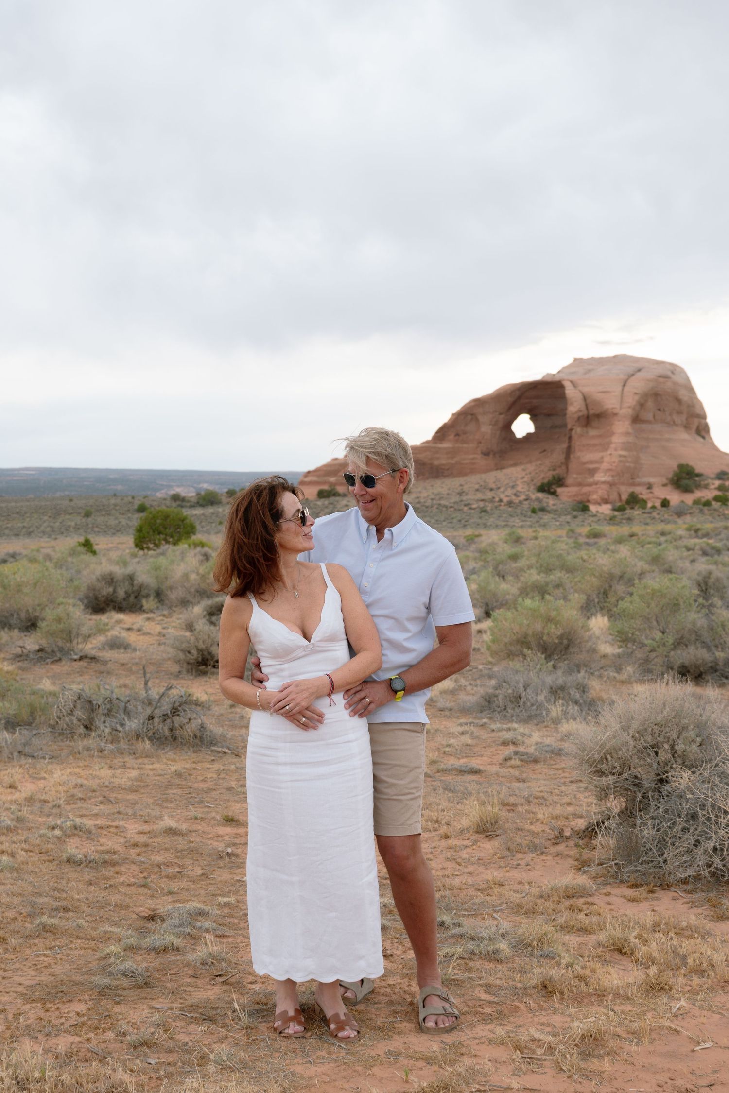 Couple in white dress and khaki shorts posing before a red rock arch formation in an arid desert landscape.