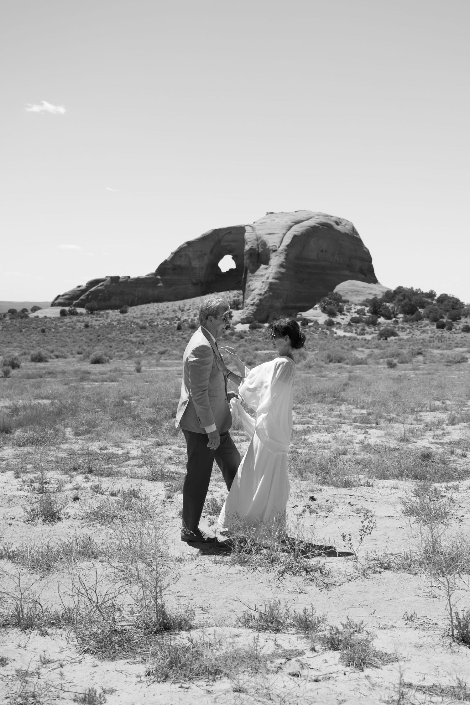 Couple holding hands in desert landscape with dramatic rock formation backdrop during outdoor wedding ceremony.