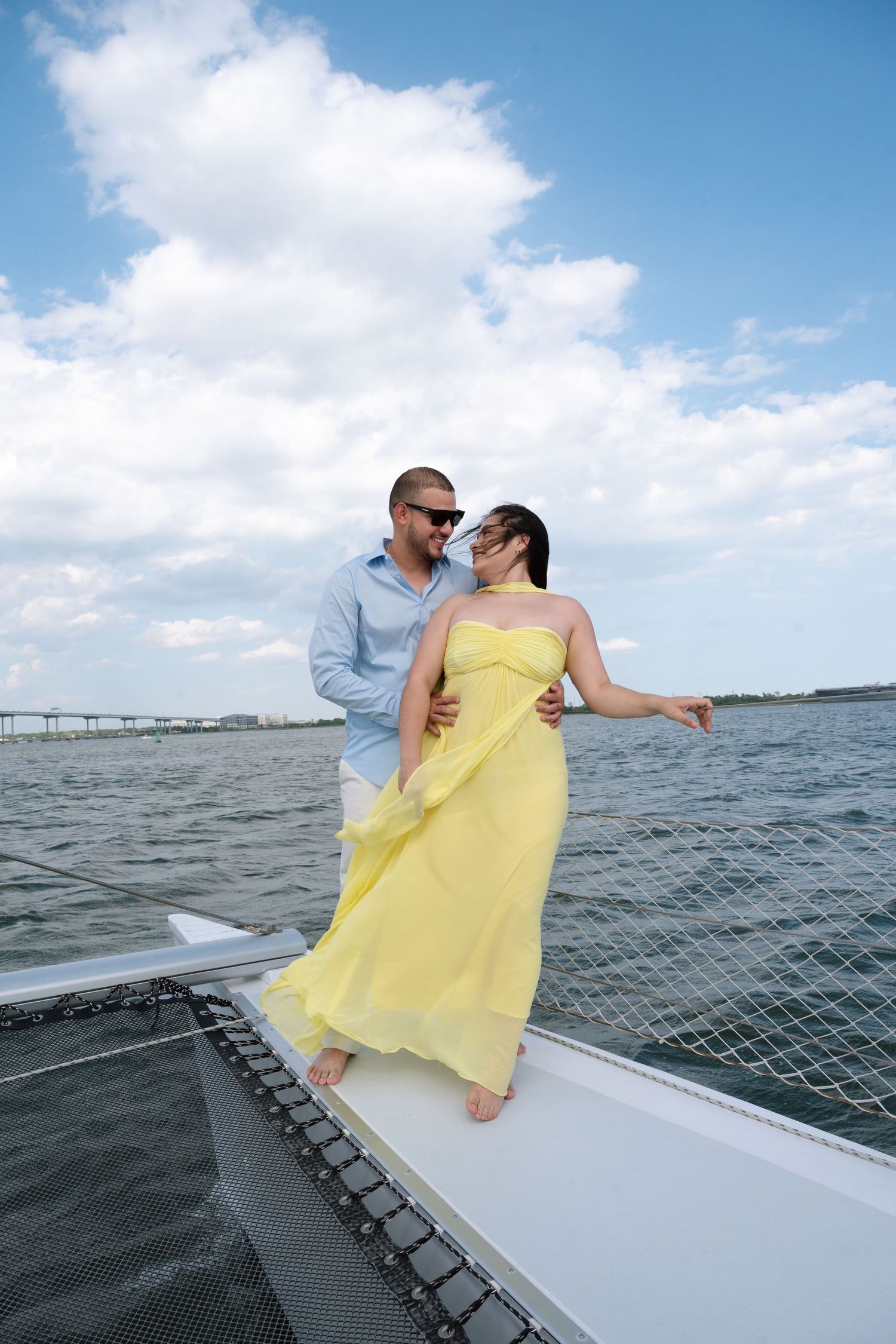 Couple in yellow dress and blue shirt embracing on sailboat bow with ocean and cloudy sky backdrop.