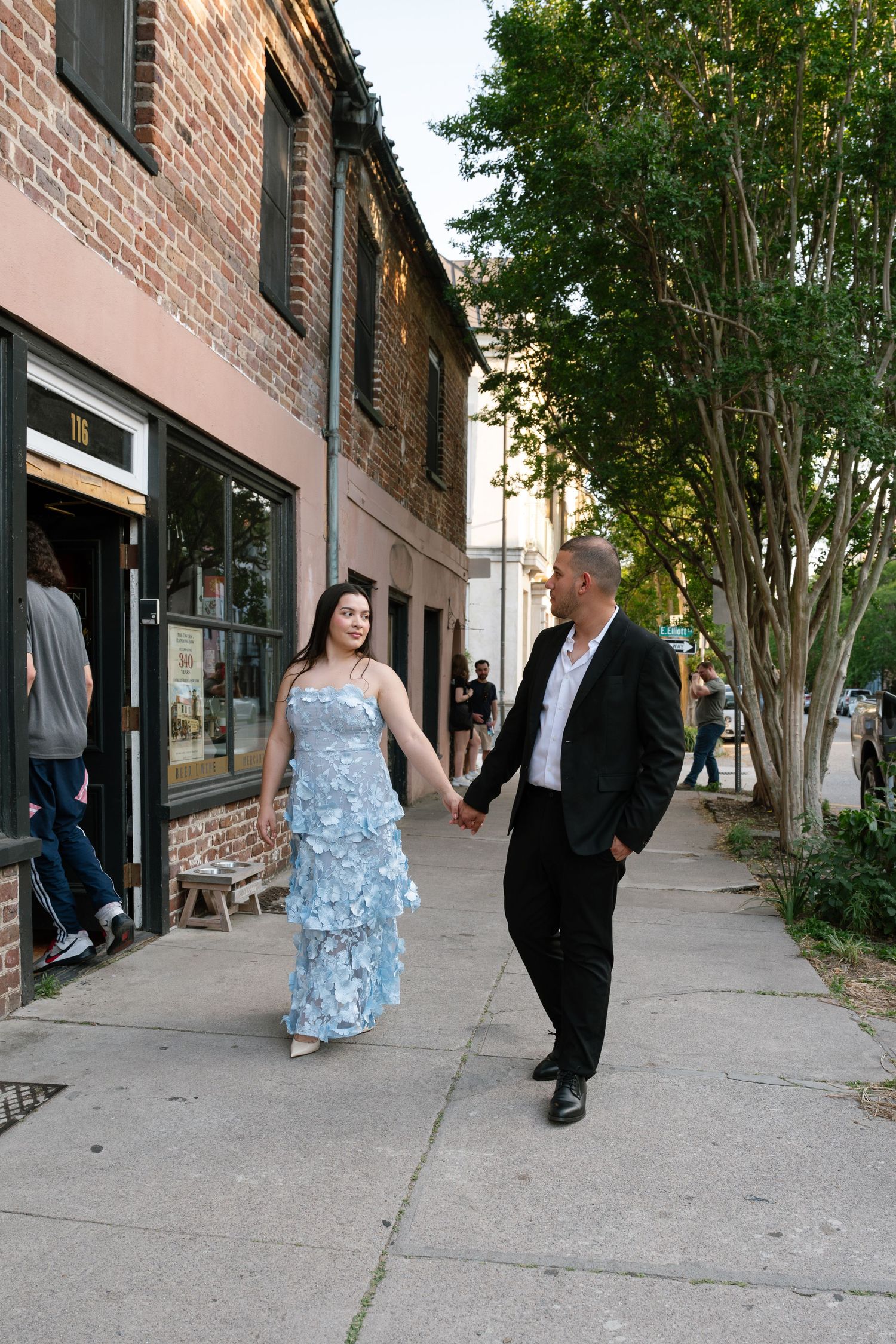 Couple holding hands on a sidewalk, woman in blue floral gown and man in black suit, walking past brick buildings.