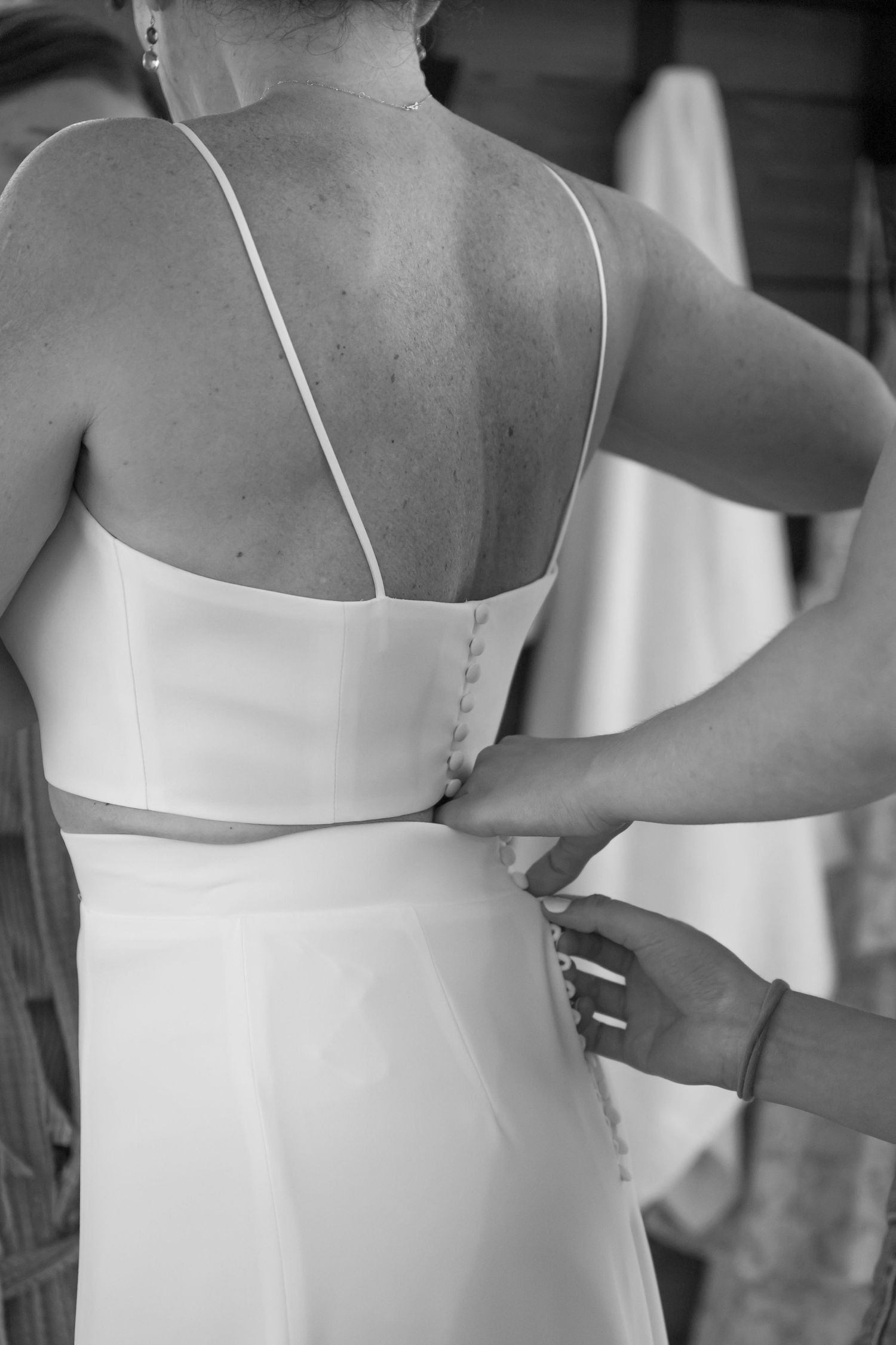 Black and white photo of a bride having her wedding dress buttoned up from behind by a helper.