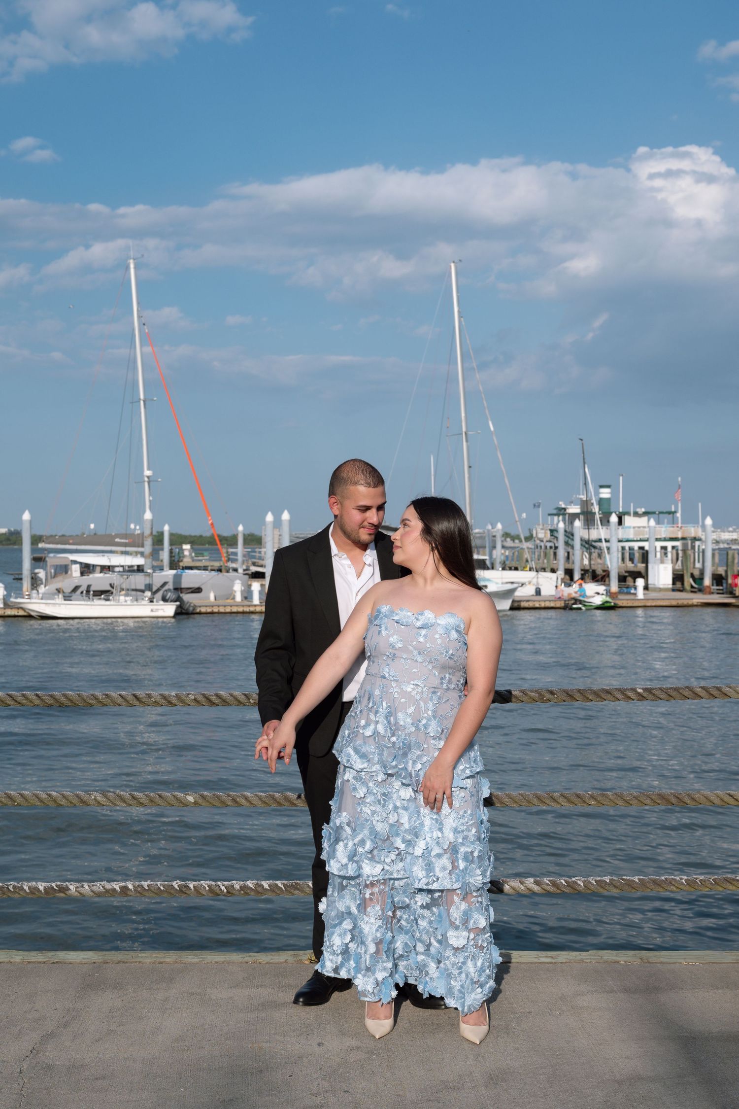 Elegant couple posing at a marina, woman in blue floral gown and man in black suit with sailboats behind.