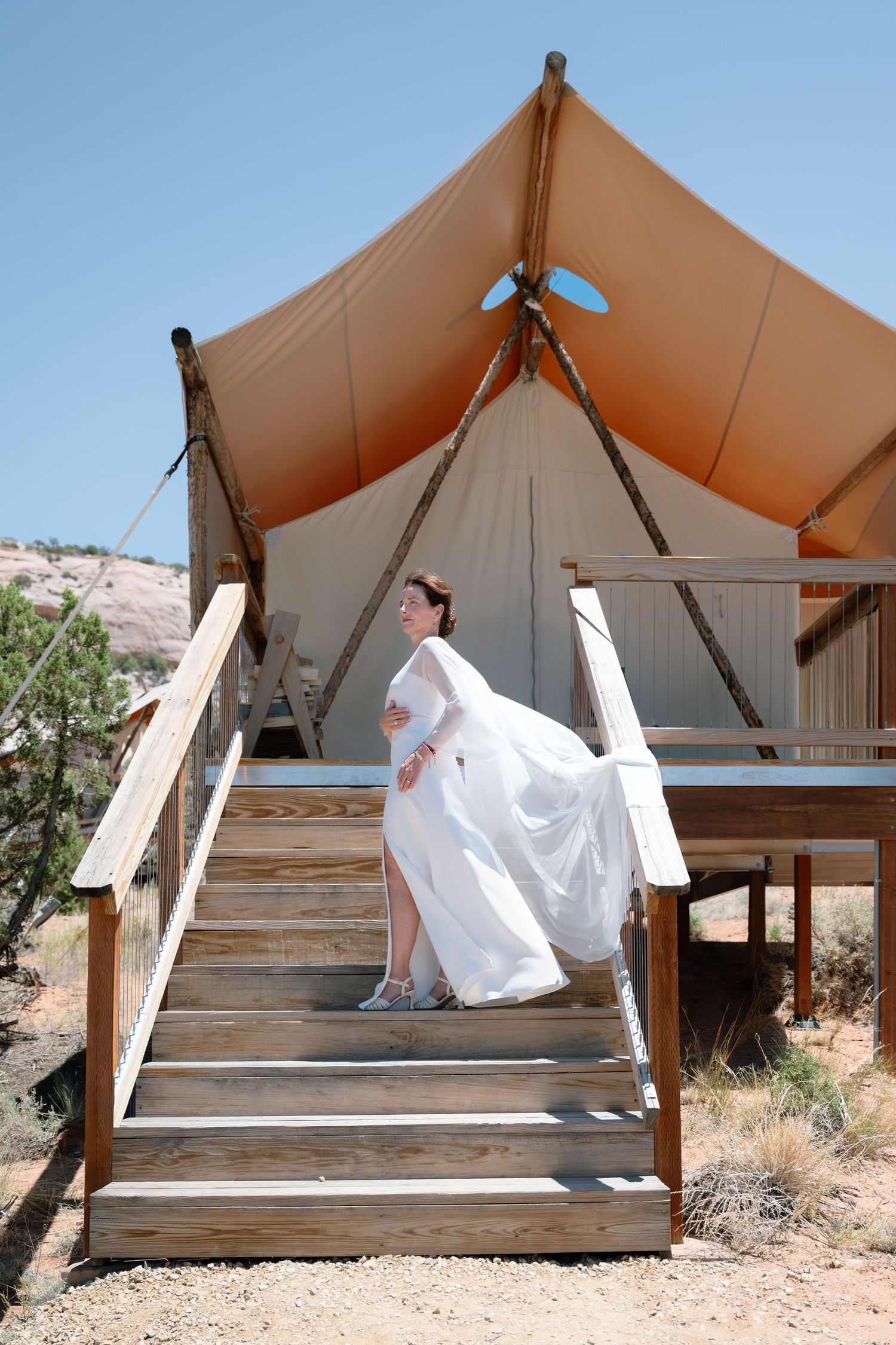Bride in white gown ascending wooden stairs of a luxury glamping tent in a desert landscape.