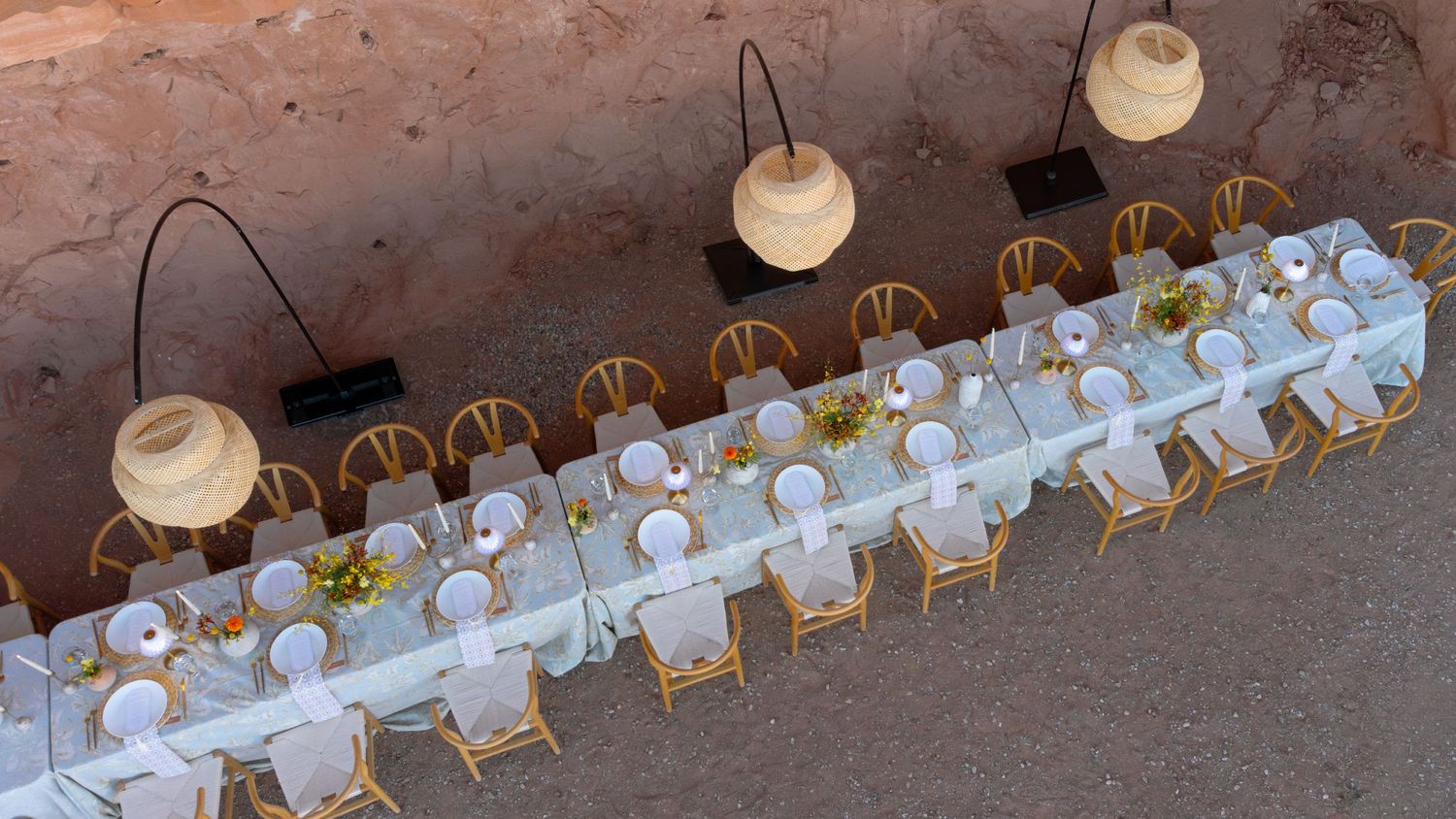 Aerial view of an elegant outdoor long dining table set with white linens, gold chairs, and wicker pendant lights in a desert.