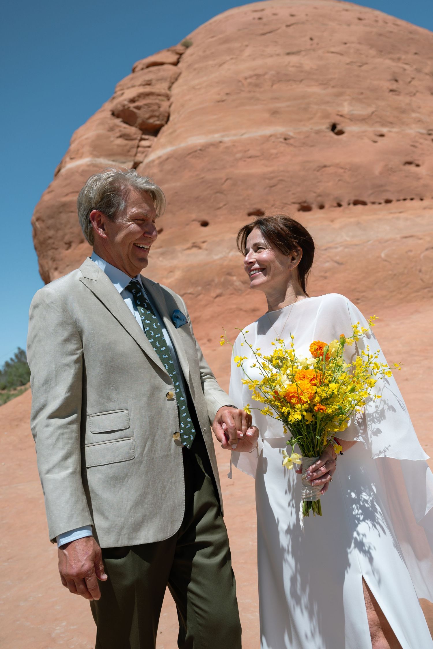 Smiling bride and groom holding hands with yellow bouquet at red rocks.