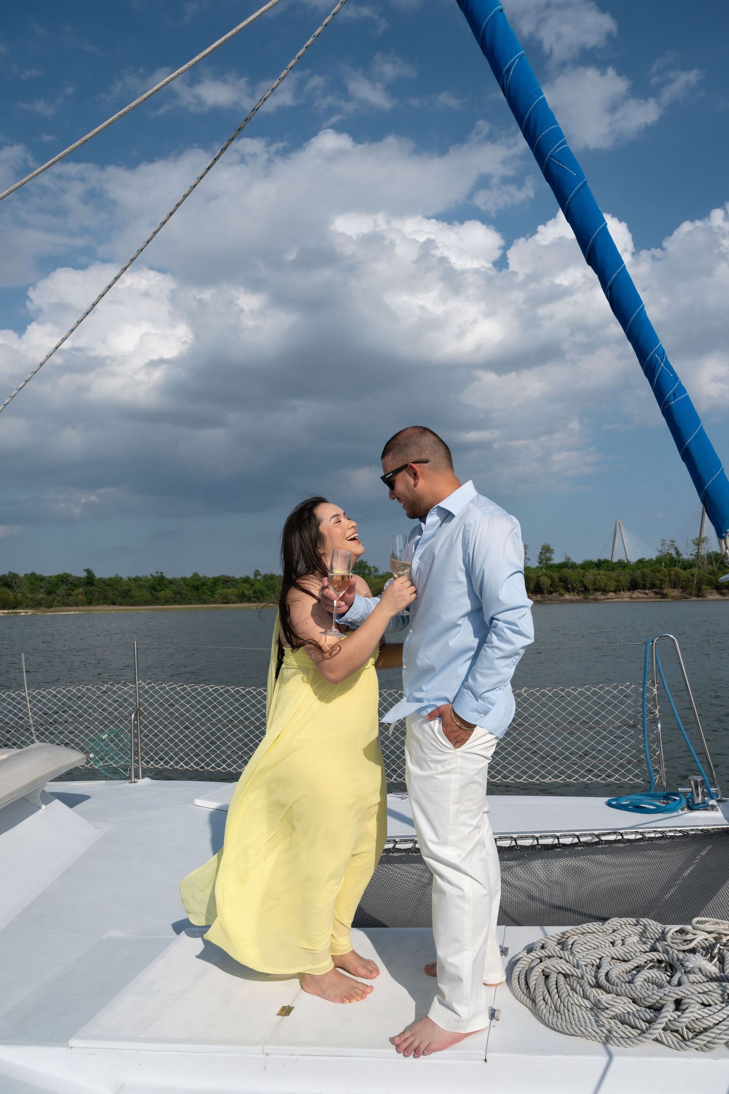 Couple celebrating on a sailboat, woman in yellow dress and man in blue shirt toasting with champagne glasses on sunny day.